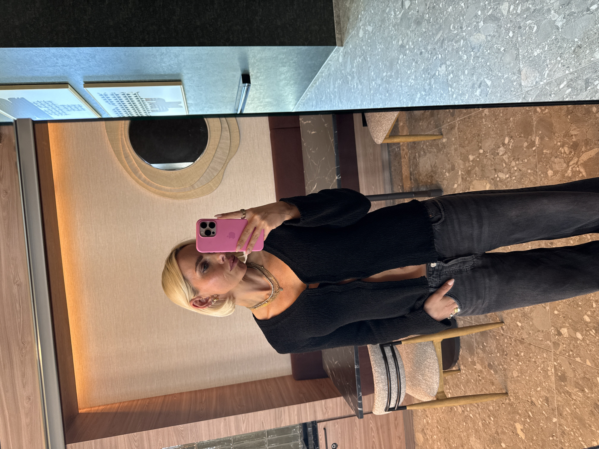 A blonde woman is captured in a mirror selfie, holding a pink-cased smartphone in her right hand to take the photo. She is looking directly at her phone, which is partially obscuring her face. She wears a black, long-sleeved, ribbed V-neck top and dark grey jeans, with the top slightly open revealing her midriff. Multiple gold-toned necklaces are layered around her neck, and she has earrings and rings on her fingers.
The setting appears to be an upscale indoor space, possibly a hotel lobby, restaurant, or modern lounge, consistent with the San Diego location. The mirror reflects a well-decorated interior. Behind the woman's reflection, there is a wall with a textured, layered round mirror and two framed artworks that depict grid-like patterns, possibly architectural drawings or cityscapes. To the left of her reflection, wooden wall paneling is visible.
In the real space, outside the mirror's reflection, a dark countertop or wall is at the very top. Below that, a light grey speckled floor or countertop is visible, transitioning to a brown speckled floor. A modern chair with a light-colored upholstered seat, black frame, and wooden legs is partially seen in the lower right, and another similar chair is hinted at. The lighting suggests an indoor setting with artificial light. The Apple logo is visible on her phone case.