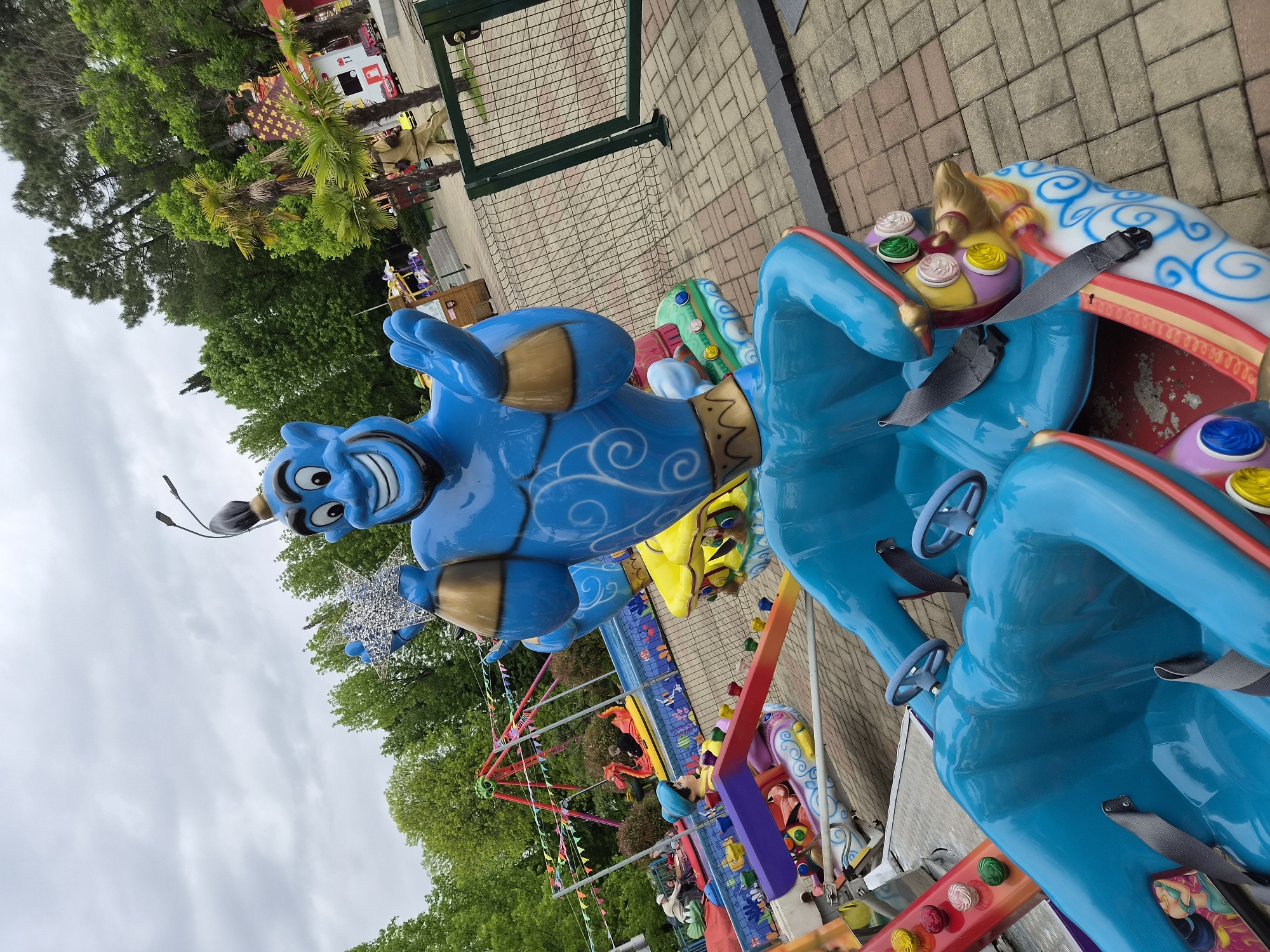 The image shows a fairground ride themed after the Genie from Aladdin, with blue fiberglass figures attached to seating areas for children. The location is Sochi, Russia, evidenced by the park's style, which is a mix of fairy tale and cartoon characters, often found in Russian and former Soviet Union parks. The seats are equipped with steering wheels and seatbelts. In the background are trees and part of a park, including a swing carousel. The sky is overcast. The image is taken at an angle looking up at the ride.