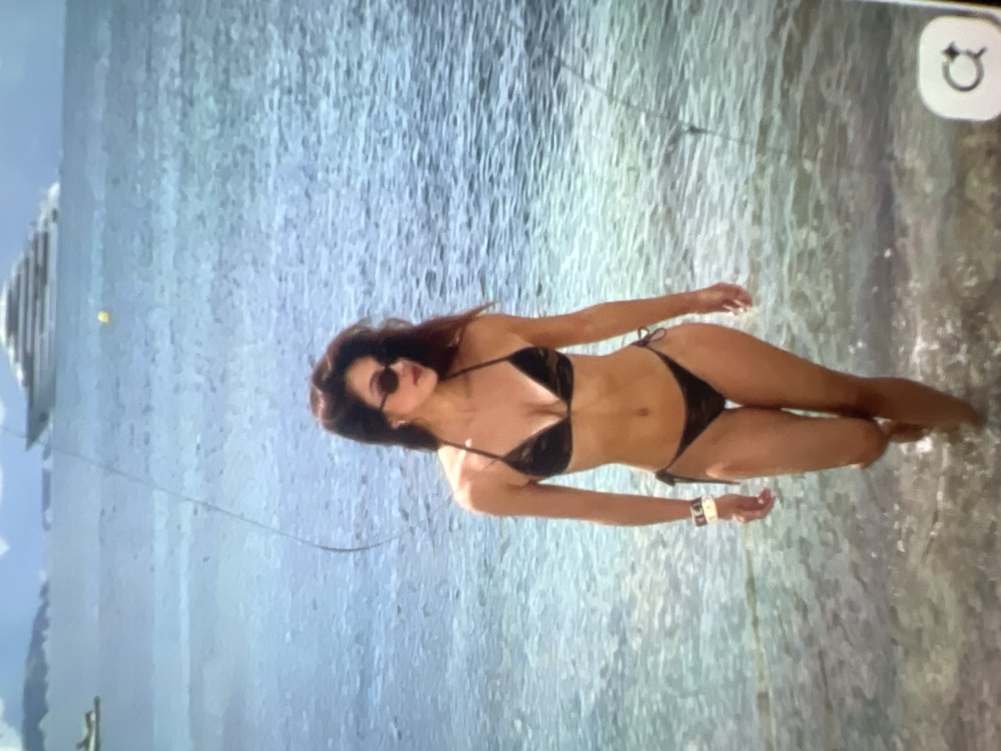 A woman in a black bikini walks in the shallow ocean water. The woman has dark hair and is wearing sunglasses. She has a white bracelet on her left wrist. In the background, a large white cruise ship is visible on the horizon, as well as distant land with mountains. The water is clear and the seabed is visible. A small boat is also visible in the far distance. The overall scene appears to be a sunny day at a beach or coastal area. The location is not Kara-Dıykan, Kyrgyzstan, as the geography does not match.