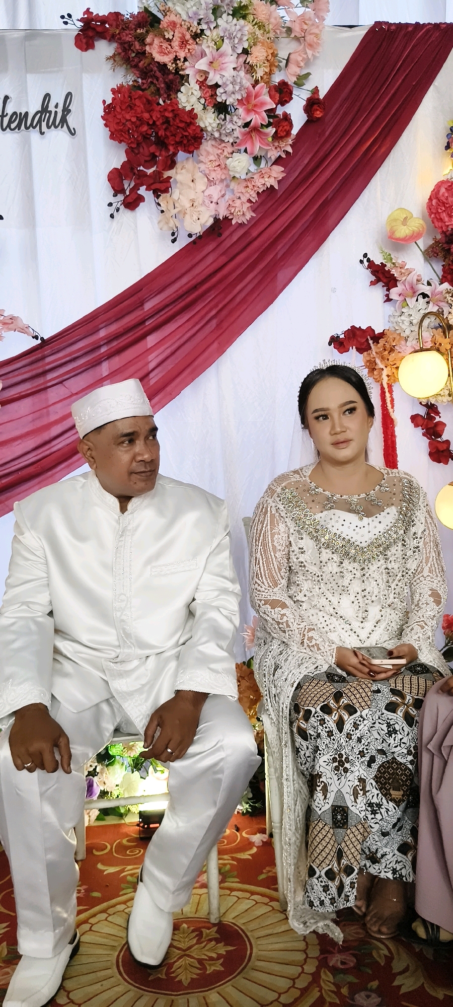 A medium shot shows a man and a woman seated at a wedding ceremony. The man, on the left, wears a white traditional suit and cap, looking to his right. The woman, on the right, wears a white wedding dress with intricate beading and a tiara. She holds a small phone in her hand and looks forward with a neutral expression. They are seated on chairs in front of a backdrop of white fabric, adorned with red drapery and floral arrangements. The floor is covered with a red carpet featuring an ornate pattern. The scene appears to be a wedding event, likely taking place in a venue in South Tangerang, Indonesia, given the traditional attire and wedding setting.