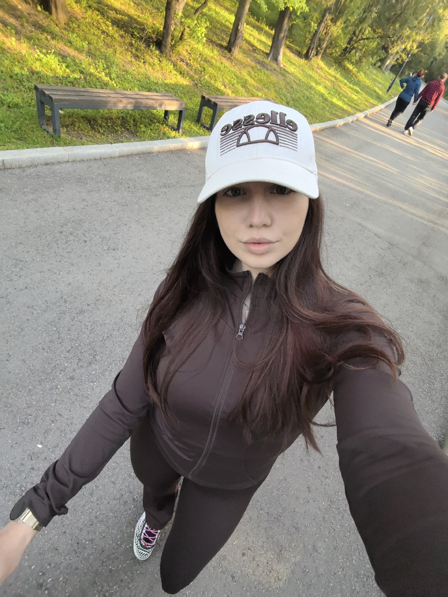 A young woman is featured in a selfie, looking directly at the camera with a neutral, calm expression. She has long, dark brown hair that falls over her shoulders and is wearing a white baseball cap. The cap prominently displays the brand name "Ellesse" in a stylized, mirrored font ("səlləE") with a distinctive arched logo in brown embroidery. She is dressed in a dark brown (possibly deep maroon or chocolate brown) athletic outfit, consisting of a zip-up jacket and matching leggings. On her left wrist, she wears a watch with a gold-colored mesh band. Her sneakers are white with black stripes and striking pink laces.
The scene is an outdoor park or green space with a paved asphalt path. To the left of the woman, on a slightly elevated grassy slope, are two dark wooden park benches with metal frames. The background is filled with lush green grass and numerous trees. The lighting is warm and golden, suggesting either late afternoon or early morning. Further down the path, two other individuals are visible walking away from the camera; one wears a blue jacket and dark pants, and the other is in a red plaid shirt and dark pants. The overall atmosphere is serene, portraying a pleasant day for outdoor activity in a public park, consistent with a location such as Stara Zagora, Bulgaria.
