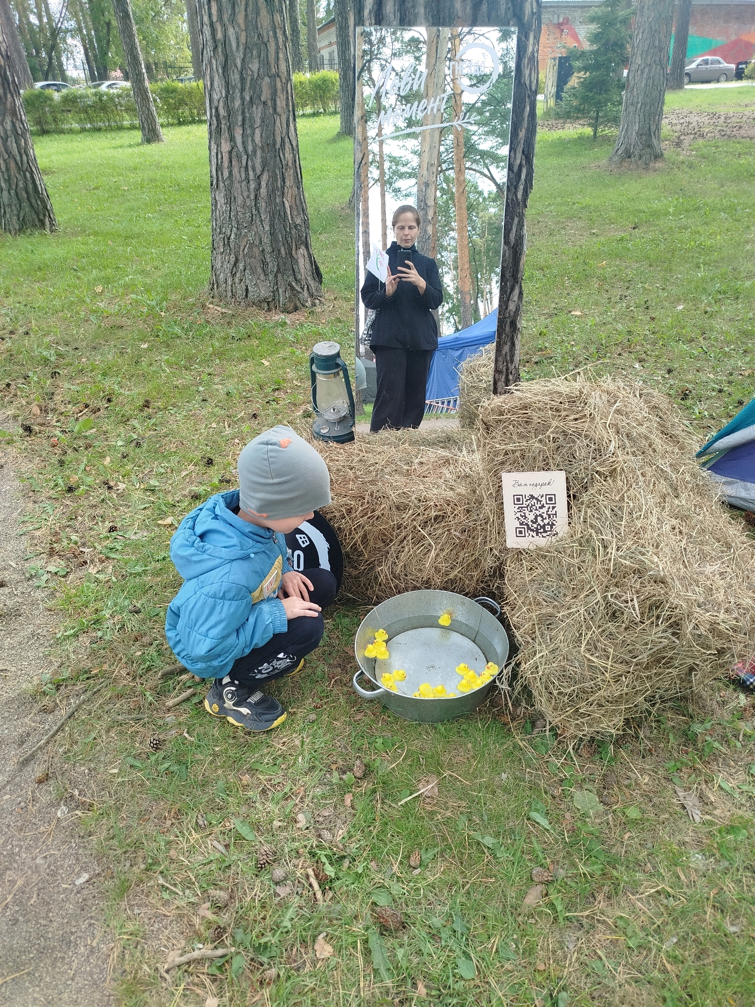 In the city of Zavodoukovsk, Russia, a young boy, wearing a blue jacket, grey hat, and dark pants with yellow accents on his shoes, is crouching on a grassy area. He appears to be interacting with a metal basin filled with water and numerous yellow rubber ducks. Behind him, a large haystack is situated next to a tall, framed mirror which reflects a woman in dark clothing holding a smartphone and a small white object. A traditional kerosene lantern stands near the haystack. A blue tent is partially visible in the background. The ground is covered with grass, scattered pinecones, and dry leaves. The overall setting suggests an outdoor recreational space, possibly a park or event area.