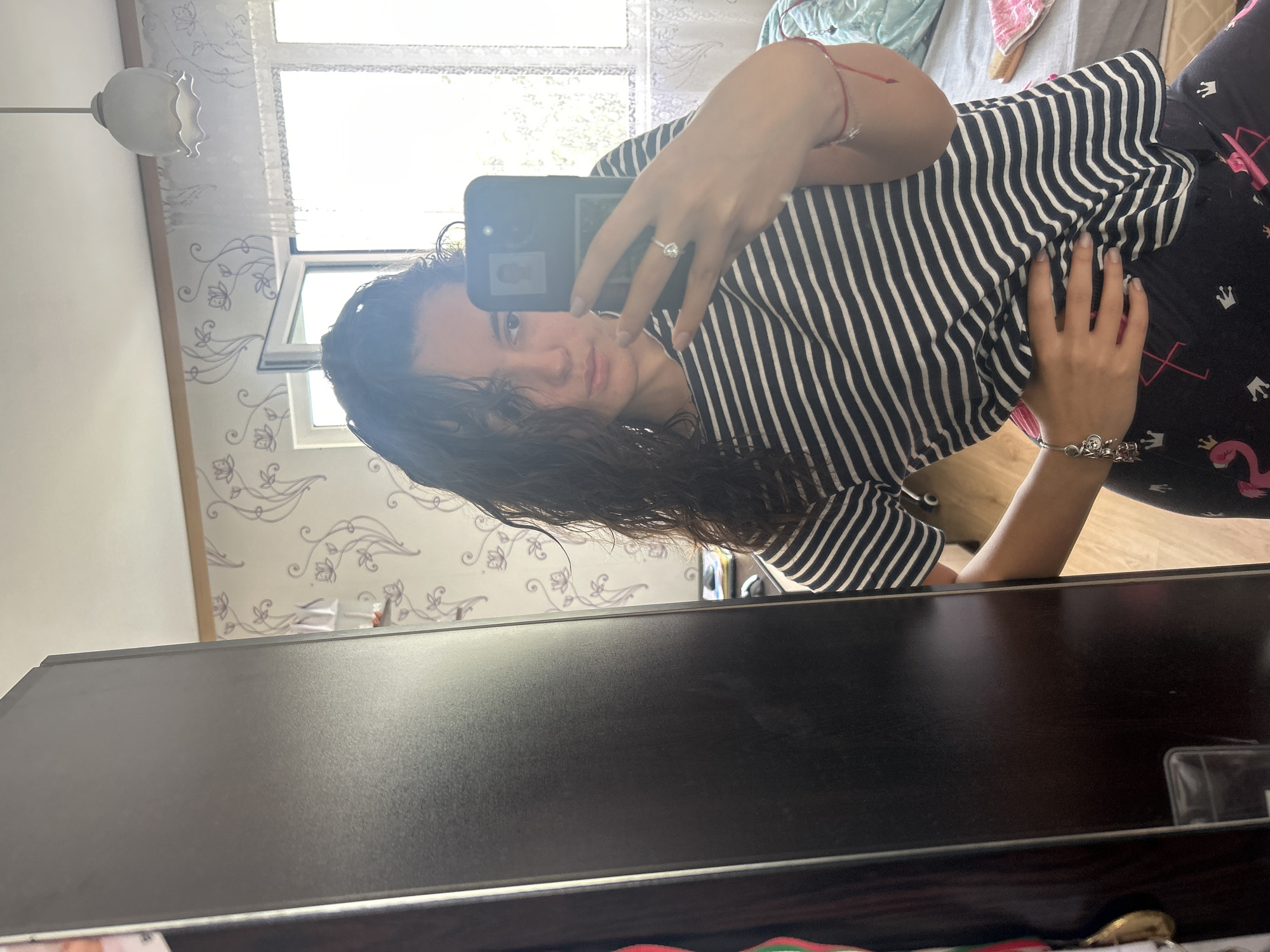 A person is taking a selfie in a bedroom. The person is wearing a black and white striped shirt and black patterned pants, and has wet dark curly hair. They are holding a black smartphone in their left hand to take the picture. The background shows a bed with a white and blue blanket, a window with white curtains, and patterned wallpaper. A ceiling light fixture is also visible. The location is the bedroom of a residence in an unknown city.