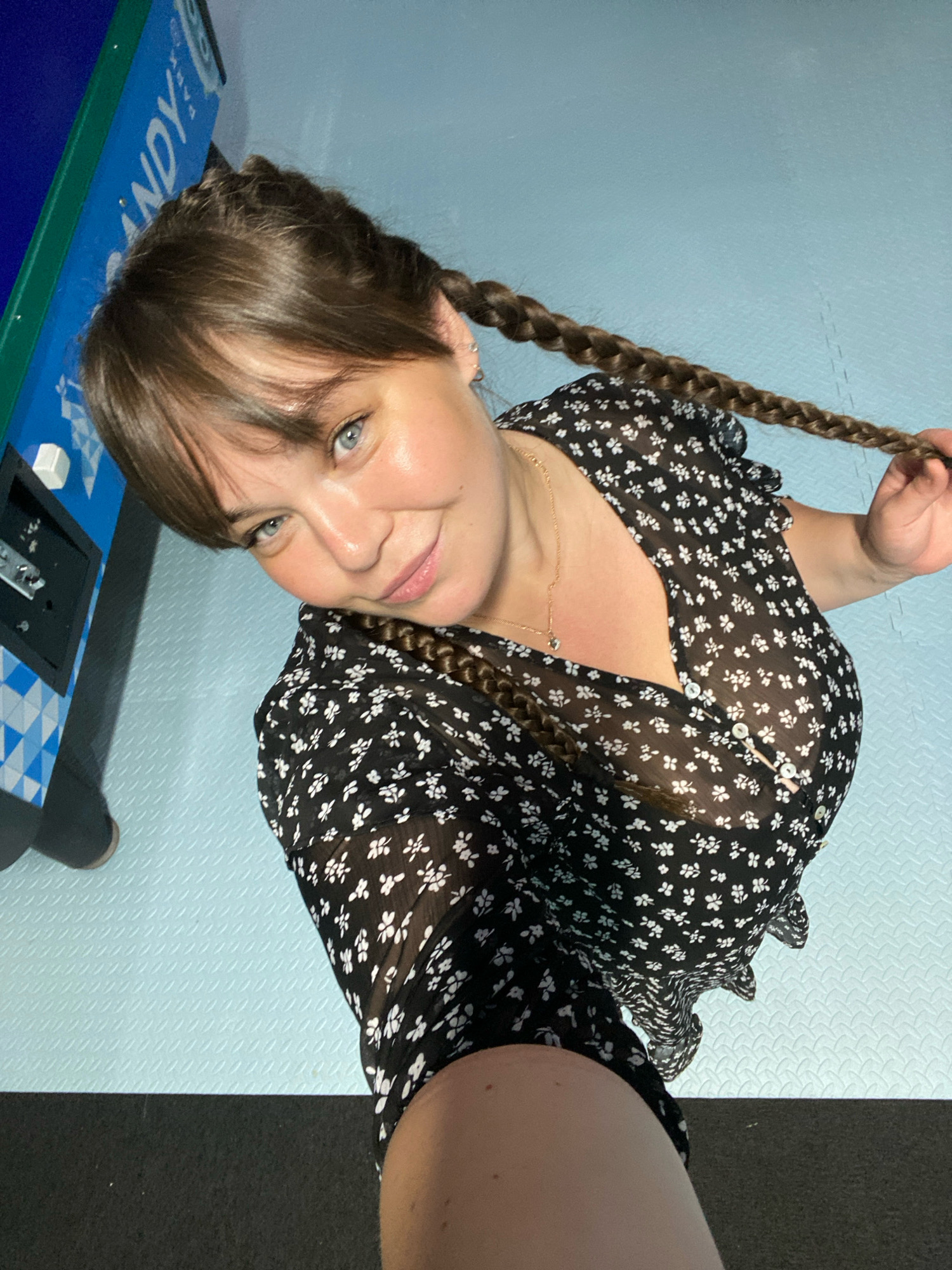 A woman with brown hair in pigtails and blue eyes is posing for a selfie. She is wearing a black dress with white flowers. She is standing next to what appears to be a game machine, possibly an arcade game. The floor is a light blue rubber mat with a textured pattern. The background wall is also a light blue color. There is a dark grey or black surface in the foreground.