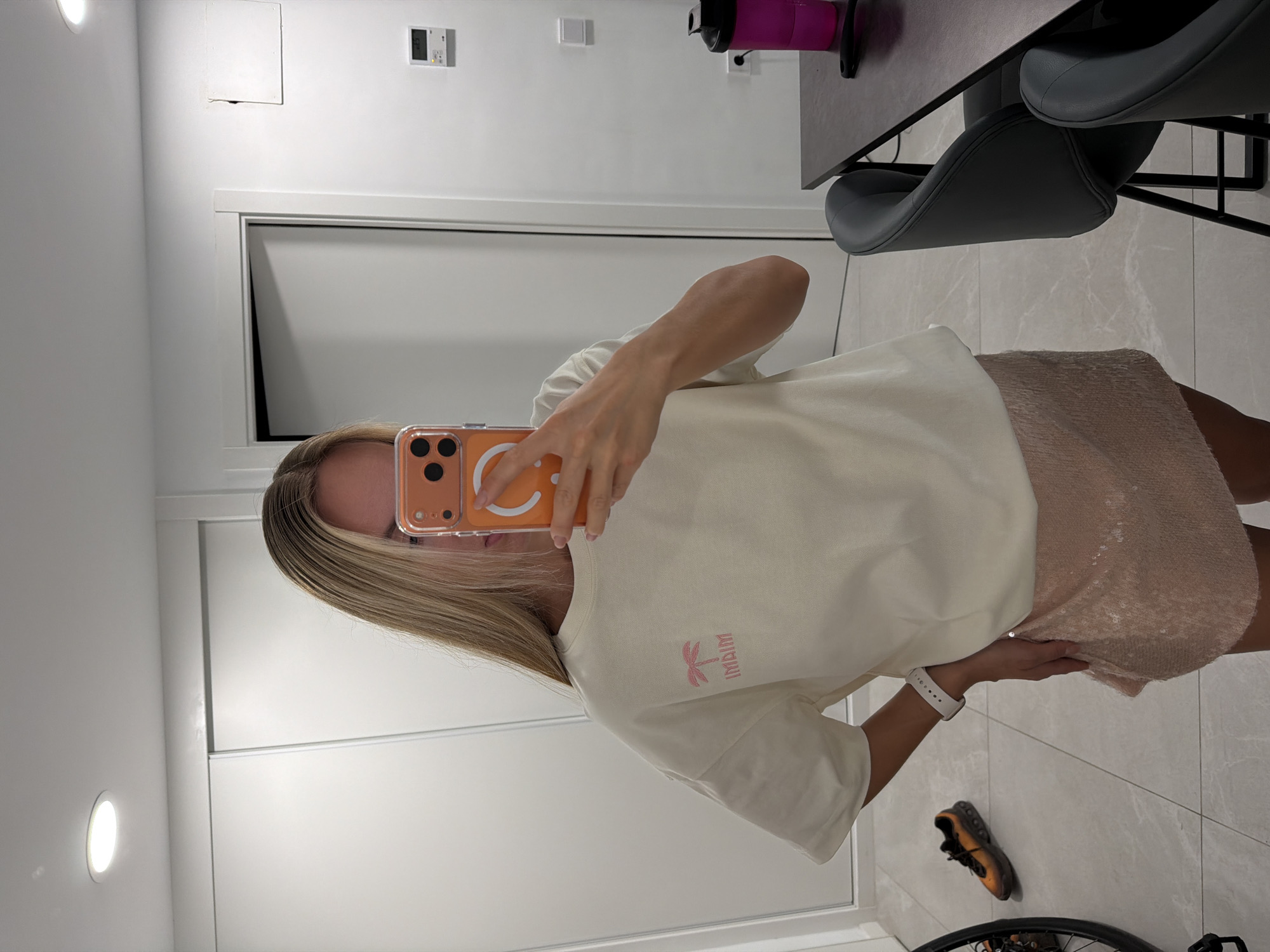 The image shows a person taking a selfie in a room with white walls and light grey tiled floors. The person has blonde hair and is wearing a light-colored t-shirt with a pink palm tree graphic and text, and a sparkling beige skirt. They are holding an orange phone case. A white watch is visible on their left wrist. In the background, there are doors, a light fixture, and a desk with a pink water bottle and a grey chair. A pair of orange and black sneakers are on the floor. The room appears to be indoors. There is no indication of the time of day or weather. The text on the t-shirt appears to read "MAIM".