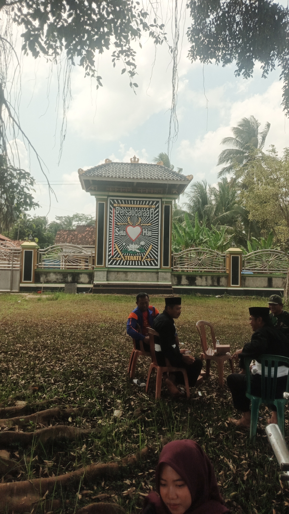 In Nabang Baru, Indonesia, an outdoor gathering takes place near a distinctive monument adorned with a heart emblem and Indonesian text. Several men are seated on chairs, engaging in conversation. The environment is characterized by dry, leafy ground, exposed tree roots, and lush green vegetation, including banana trees, in the background. The sky is partly cloudy, with palm trees visible. A woman wearing a maroon hijab is present in the foreground, slightly out of focus. The monument itself is a significant focal point, suggesting a cultural or community gathering.