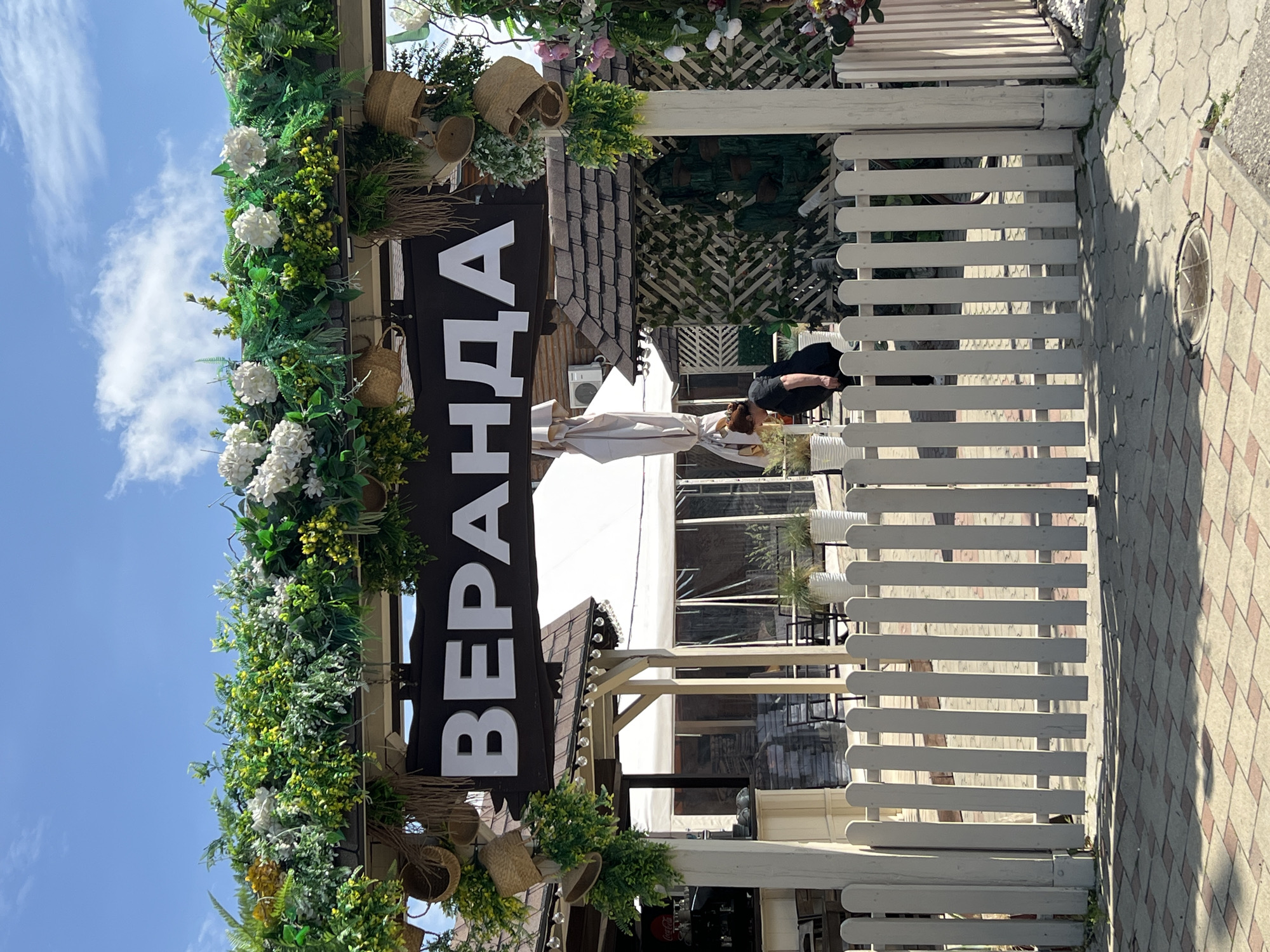 The image shows the entrance to a restaurant or cafe called "Веранда" (Veranda). The sign is vertical, with white letters on a black background, and is decorated with greenery and hanging baskets. The establishment has a white picket fence. A person is visible inside the entrance. The location is likely Krasnodar, Russia, given the Cyrillic lettering. The pavement outside the fence is patterned with tiles. The sky is blue with some clouds.