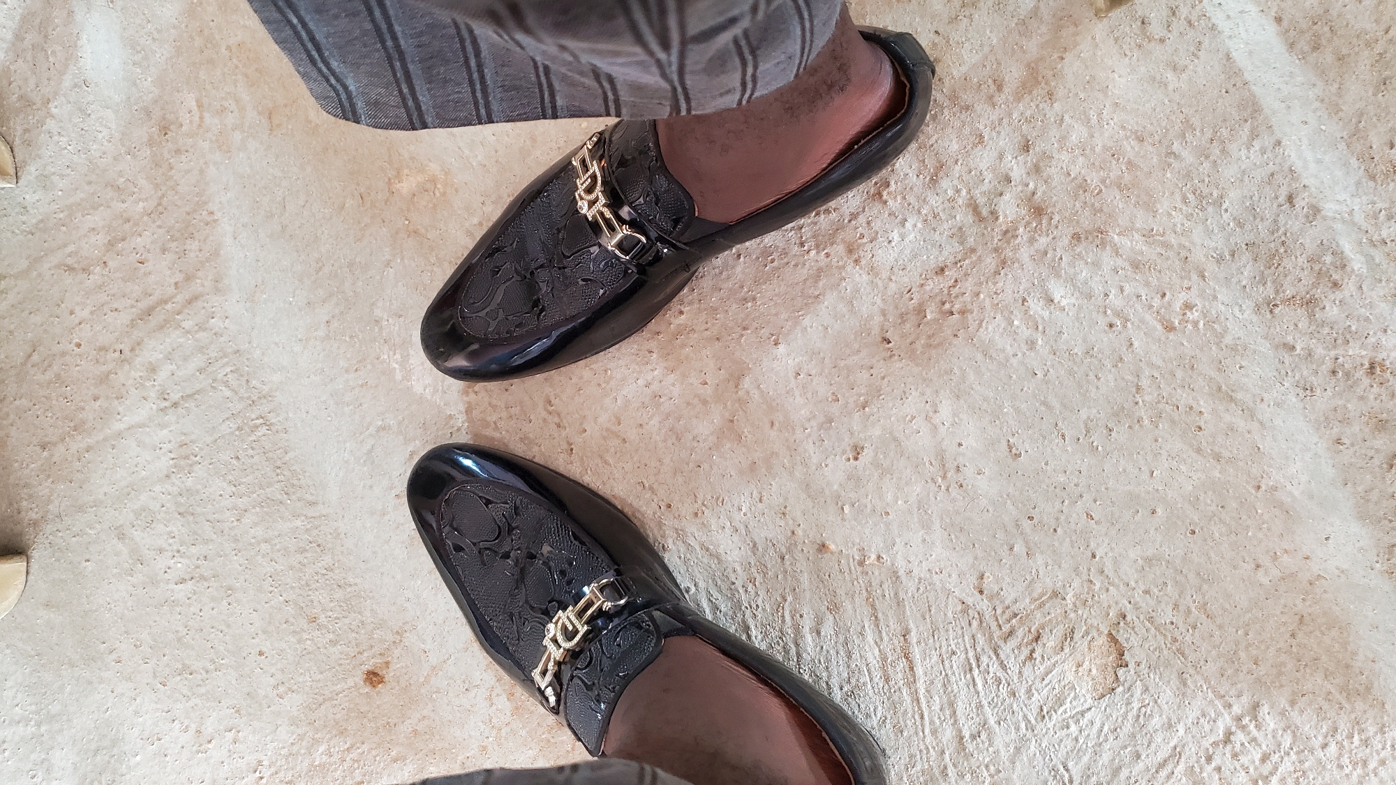 The image shows a pair of black and brown leather loafers with a gold chain detail on the vamp. The shoes are placed on a light-colored, textured surface that appears to be a floor. The right foot is partially visible, wearing a gray pinstriped pant leg. The scene is likely indoors or on a paved outdoor surface. The location is identified as Jalingo, Nigeria, although there are no specific visual cues in the image to confirm this. The lighting is even, suggesting it's daytime.