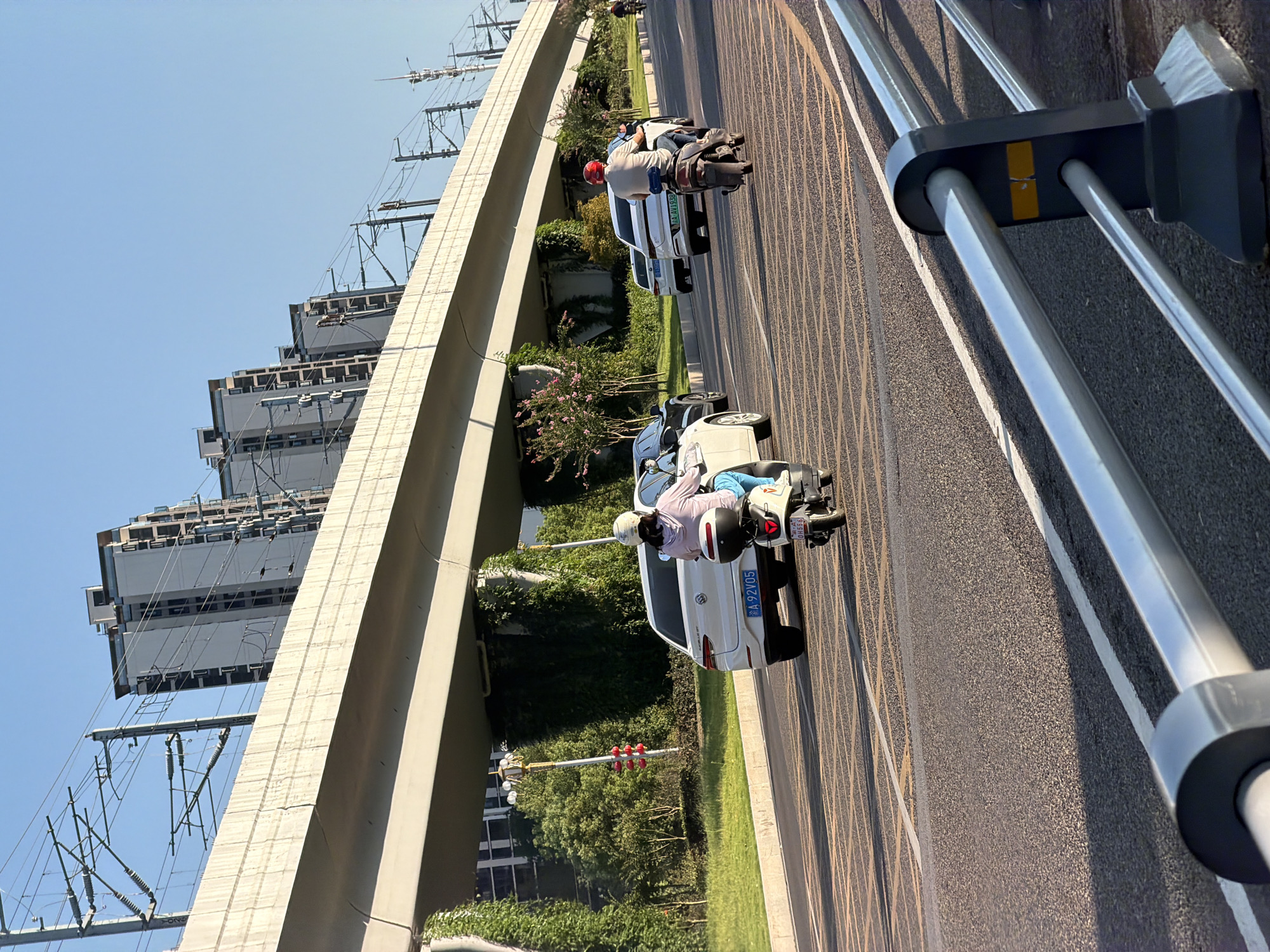 In Jiangjin, China, under a clear blue sky, an elevated highway or overpass looms over a road. Several modern residential buildings are visible in the background, interspersed with power lines and poles. On the road below, a white car with license plate "AA 92V05" is being followed by a person on a white scooter wearing a helmet and a light pink jacket. Further down the road, another person on a darker scooter with a red helmet is visible. The road has clear lane markings and a guardrail in the foreground. The scene appears to be set in an urban or suburban area during daylight.