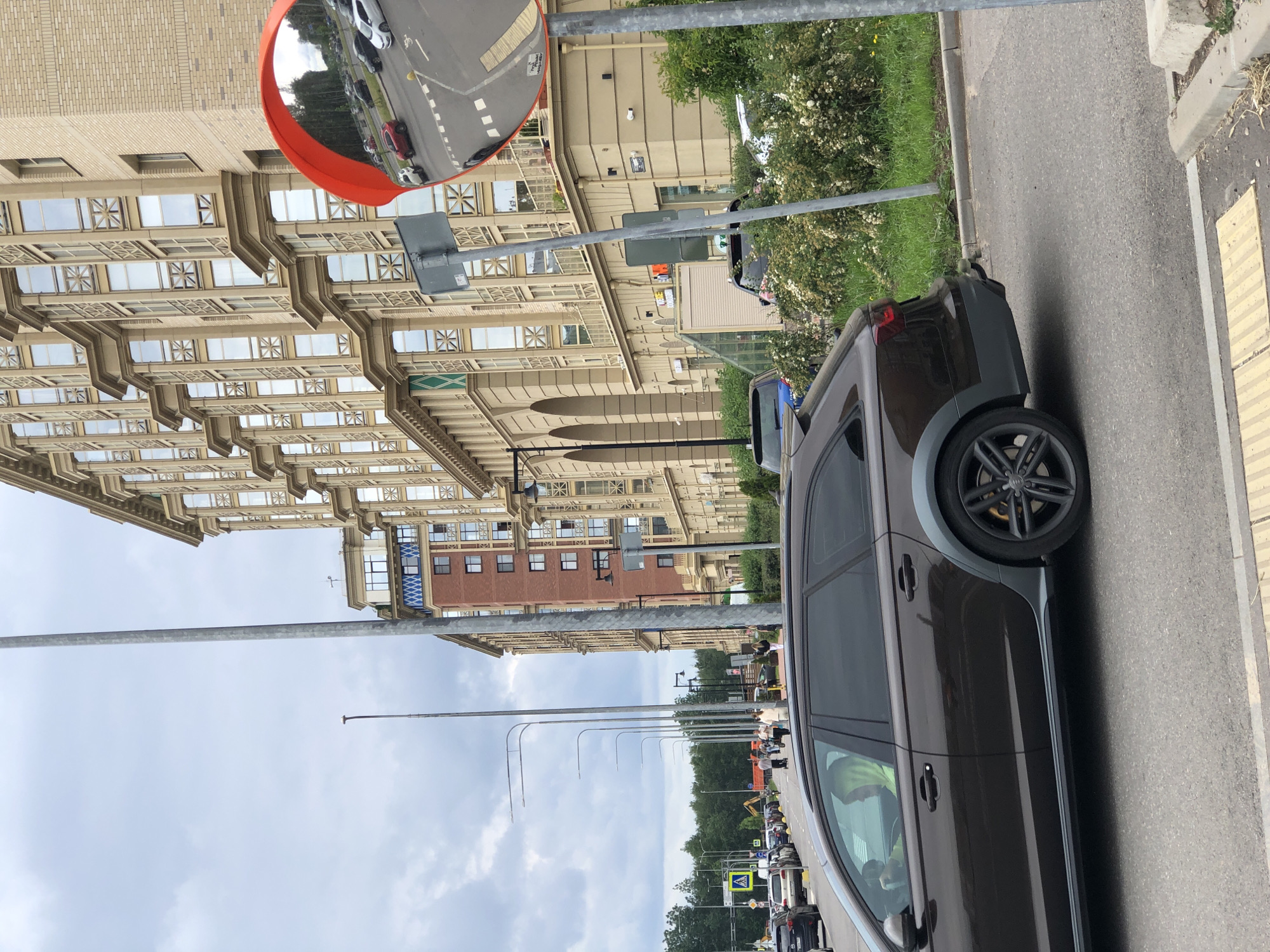 The image shows a street scene in Saint Petersburg, Russia. In the foreground, a dark brown station wagon is parked or driving on the right side of the road. A person wearing a bright green hooded sweatshirt is visible in the driver's seat. Across the street, a modern, multi-story building with a beige facade and numerous windows dominates the background. To the right of the building, a large, round convex mirror is mounted on a pole, reflecting traffic and the surrounding environment. Several other cars are visible on the street and in the background, as well as trees and a cloudy sky. Pedestrians are also present, walking on the sidewalk in the middle ground. The street appears to be an urban or suburban area, with a mix of residential and commercial architecture.