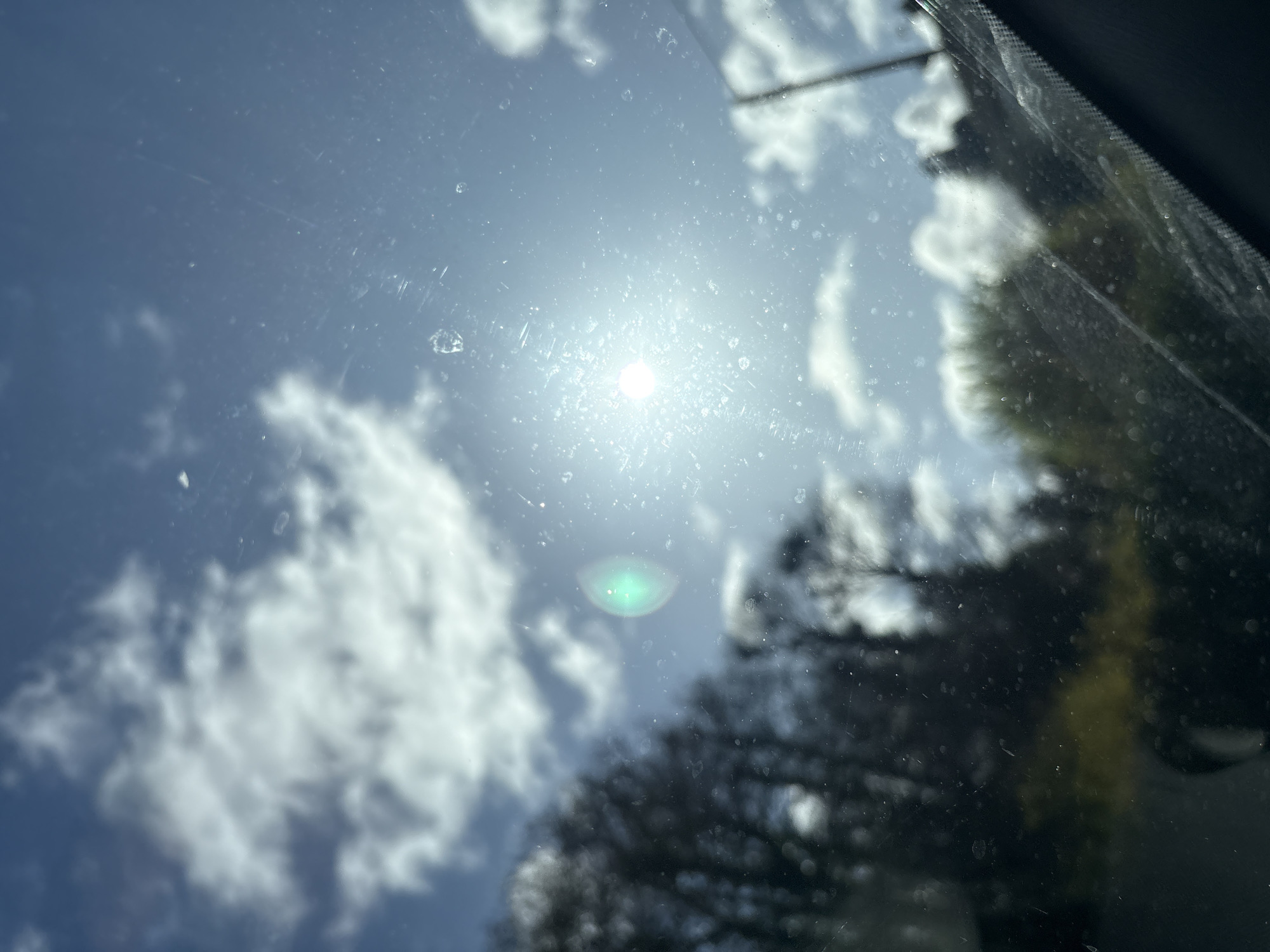 The image depicts an outdoor scene in the city of Ruyton-XI-Towns, United Kingdom, viewed through a transparent, soiled, and scratched surface, likely a vehicle's windshield. The upper and central portions reveal a bright blue sky with scattered white cumulus clouds. A radiant sun is positioned in the upper right, generating a pronounced greenish circular lens flare slightly to its left and a starburst effect around its own brilliant core. The foreground glass surface is marred by numerous small, irregular specks, consistent with dried water droplets or dust, and multiple fine linear abrasions. The mid-ground and background feature out-of-focus dark silhouettes of trees, some appearing bare and deciduous, alongside areas of lighter green foliage, primarily on the left and lower-left. A dark, vertical post, possibly a utility pole, is faintly visible on the far right. The lower right frame shows an indistinct, dark section indicative of a vehicle's interior trim. No human figures or specific interactions are present.