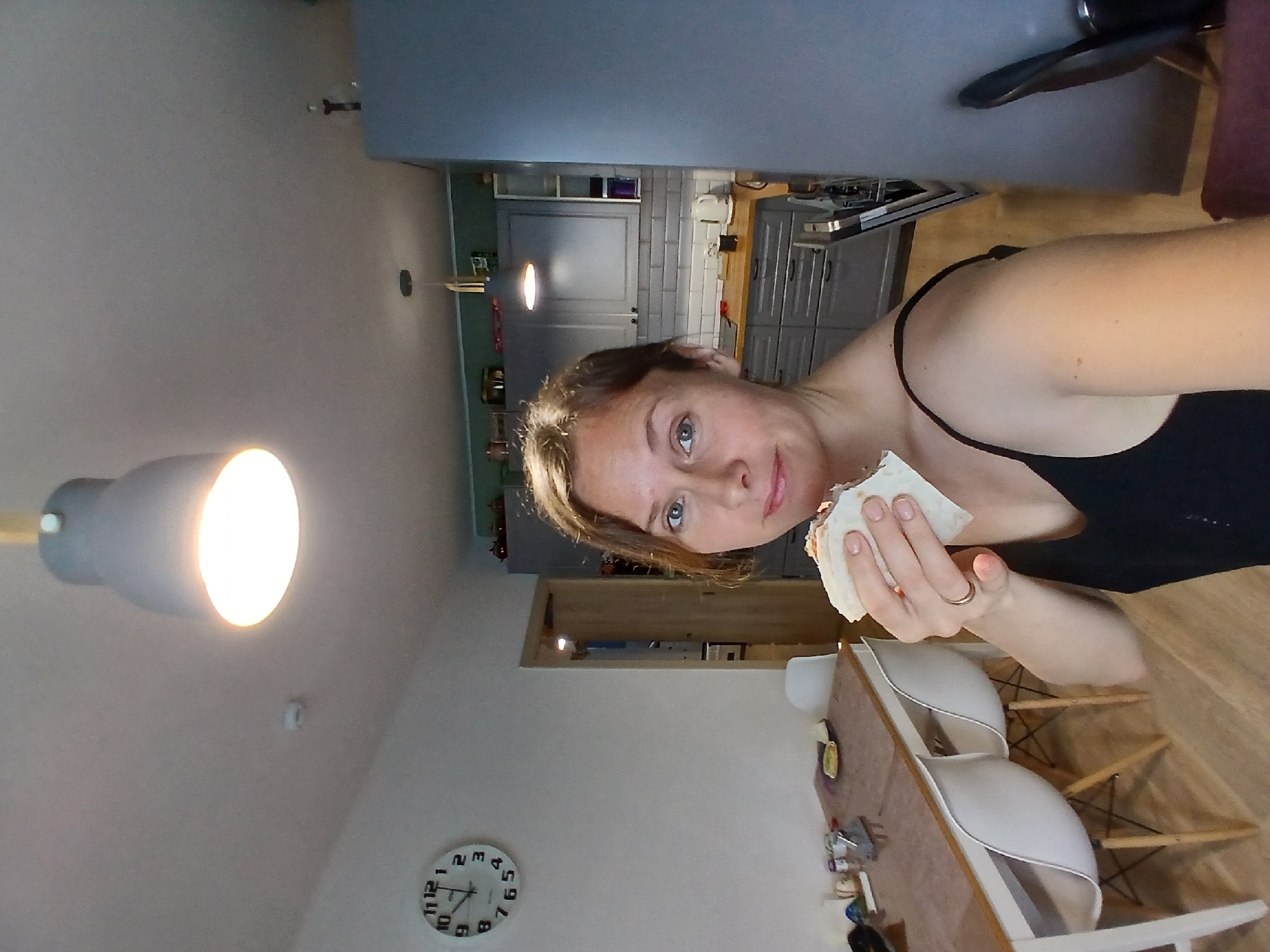 A woman is holding a sandwich to her mouth, with her right hand. She is wearing a black tank top. The woman is in a room that appears to be a kitchen and dining area. To her left, there is a dining table with white chairs. A circular clock is mounted on the wall above the table. To her right, there are kitchen cabinets and a countertop with a white kettle. The room has a light-colored ceiling with a hanging light fixture. The floor is made of light-colored wood. The image is not taken in Yekaterinburg, Russia.