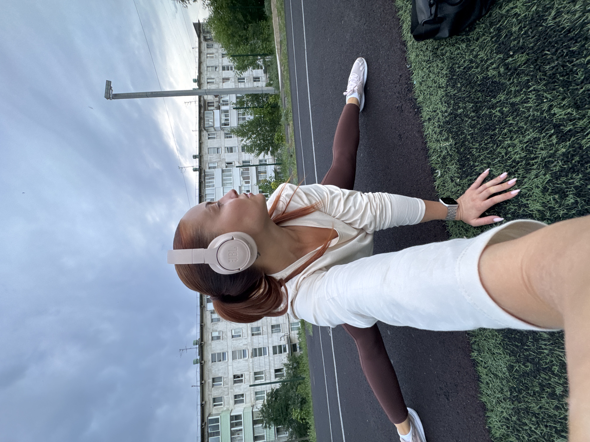A woman is doing a split on a track next to an apartment building in what appears to be the city of Chaikovsky, Russia. She is wearing headphones and athletic clothing, consisting of a white shirt and brown leggings, and white sneakers. Her hair is in a ponytail. She is using one hand to hold a phone to take the picture and uses the other to support herself on the ground. A black bag rests on the artificial grass next to the track. The weather appears overcast.