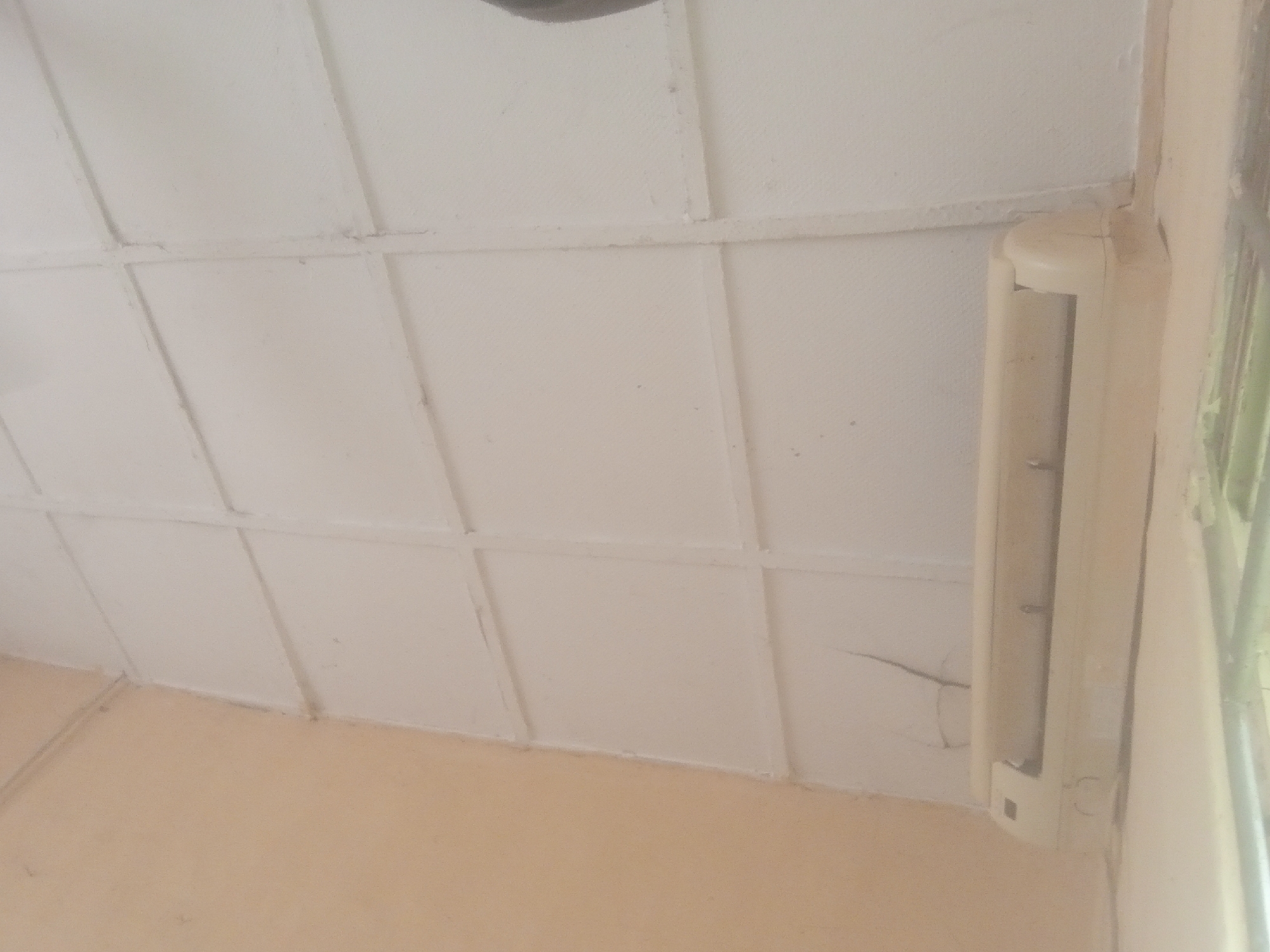 The image shows an indoor scene, likely a room with a light-colored, tiled ceiling. A beige air conditioning unit is mounted vertically on the wall on the right side of the image, adjacent to a window with a light-colored curtain. The ceiling has a grid pattern formed by white beams. The walls are painted a pale, peachy color. There are some visible cracks on the wall beneath the air conditioning unit. The lighting suggests it is daytime, but no specific time can be determined. The scene appears to be a simple interior space without any people present. There is no visible text in the image.