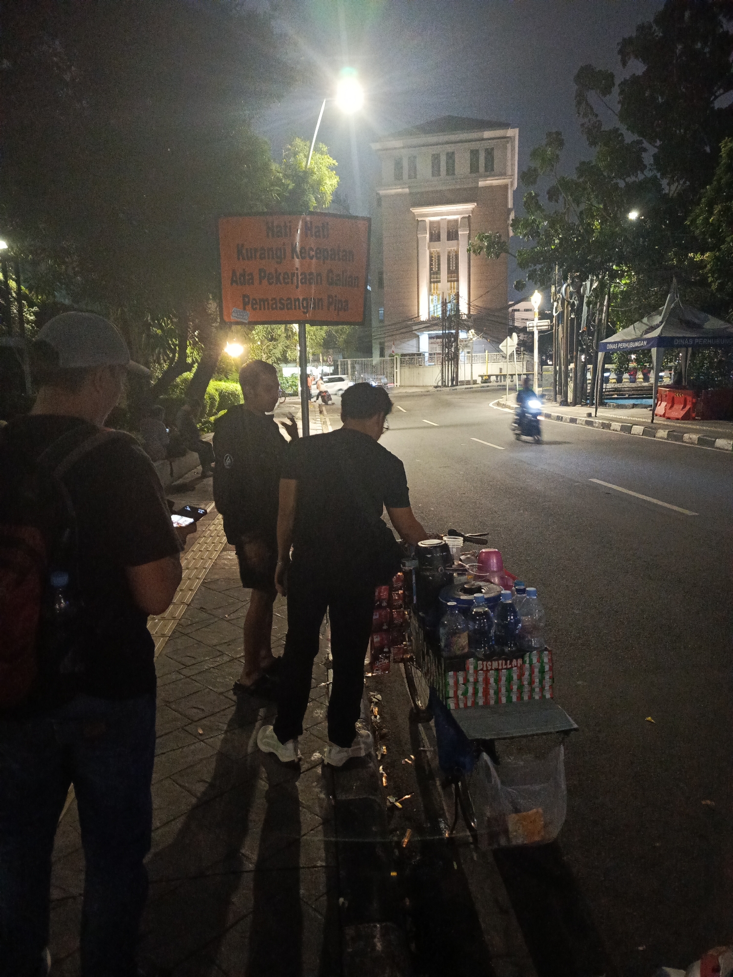 The image captures a street scene at night in Central Jakarta, Indonesia, under the glow of streetlights. In the foreground, three men stand on a tiled pedestrian walkway. The man on the left is wearing a dark t-shirt, blue jeans, and a grey baseball cap, with a red backpack visible. The central figure is wearing a dark t-shirt and shorts, looking away from the camera. The man on the right is wearing a dark t-shirt and pants, facing the street.
Adjacent to the walkway, a street vendor's cart is laden with bottled drinks, cups, and other items, covered partially by a dark tarp. The street itself is paved and appears wet, reflecting the streetlights. A motorcycle is seen in motion on the road, its headlights illuminated.
Further down the street, a tall, multi-story building with lit windows stands prominently. To the right, a blue tent with text that appears to say "DINAS PERHUBUNGAN" (Department of Transportation) is set up, flanked by red safety barriers. Trees with dense foliage line the sides of the street, adding to the urban night atmosphere. An orange traffic sign with Indonesian text, reading "Hati-Hati Kurangi Kecepatan Ada Pekerjaan Galian Pemasangan Pipa" (Be Careful Slow Down There is Pipe Installation Excavation Work), is visible near the men, indicating ongoing roadwork. The overall impression is one of a functional, active urban environment transitioning into the nighttime.
