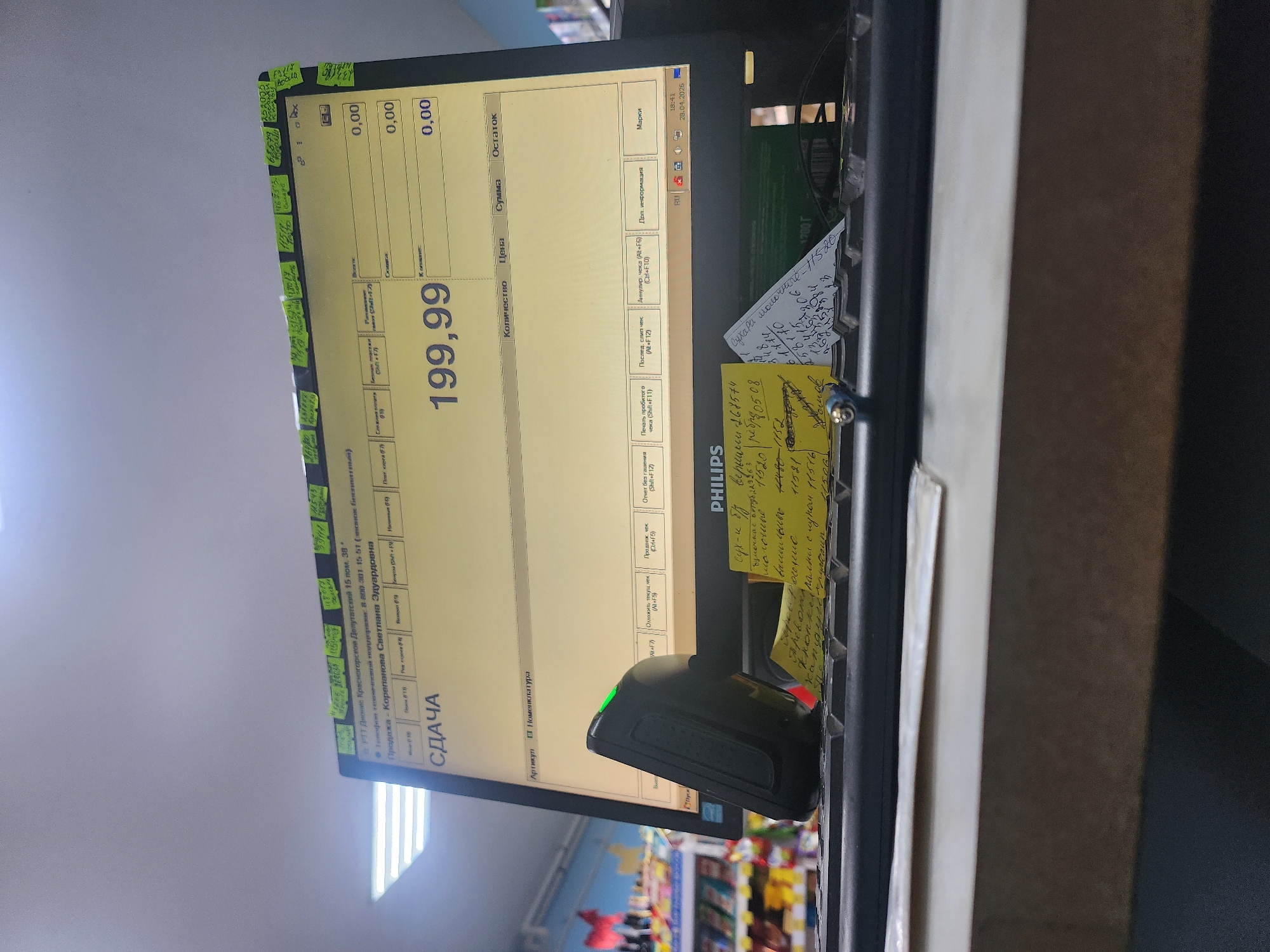 The image displays a point-of-sale (POS) system within a retail store, likely in Krasnogorskoye, Russia. The central subject is a Philips computer monitor, positioned vertically, showing a transaction interface with a prominent total of "199,99". Other fields like "Количество" (Quantity), "Сумма" (Amount), "Цена" (Price), and "Остаток" (Remainder) display "0,00". The screen also indicates "СДАЧА" (Change) and "Продажа - Корепанова Светлана Эдуардовна" (Sale - Korepanova Svetlana Eduardovna), suggesting a cashier's name.
A black barcode scanner or handheld device rests near the monitor. The system clock shows "18:41" and the date "28.04.2025". The store's address, "РТТ Дионис Красногорское Депутатский 15 пом. 38*", is visible on the interface.
Several handwritten sticky notes are affixed to the monitor's frame. These notes contain lists of items, codes, and possibly expiration dates, such as "Сухари молочные - 1/5 20." (Milk rusks - 1/5 20.), "сух-и б/у веришни 168574" (dried/used goods, code 168574), and the word "просрочение" (expired/overdue). Other numbers like "48017", "26980", "251757", "11520", and "14480-1152" are visible on the notes. The background shows blurred retail shelving with various products, confirming the store environment.