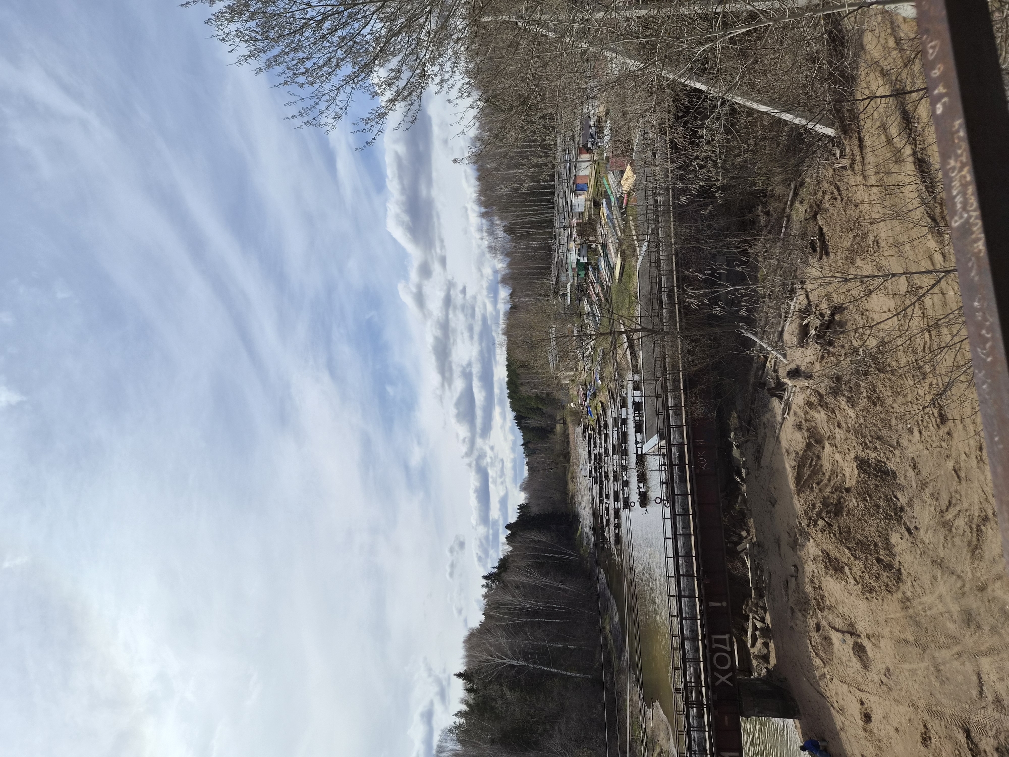 The image shows a view of a riverbank and a section of the Kama River in the city of Chaikovsky, Russia. The perspective is elevated, possibly from a bridge or high bank overlooking the river. The sky is blue with streaky clouds.
On the right side of the image is a sandy riverbank with sparse vegetation and bare trees. A metal structure, likely part of a bridge or walkway, is visible in the foreground with the inscription "ХОД" in Russian on its side.
Across the water, there is a boat launch area with numerous boats stored on racks or in slips along the shore. A mix of trees, some coniferous and some bare deciduous trees, forms a backdrop behind the boat storage area.