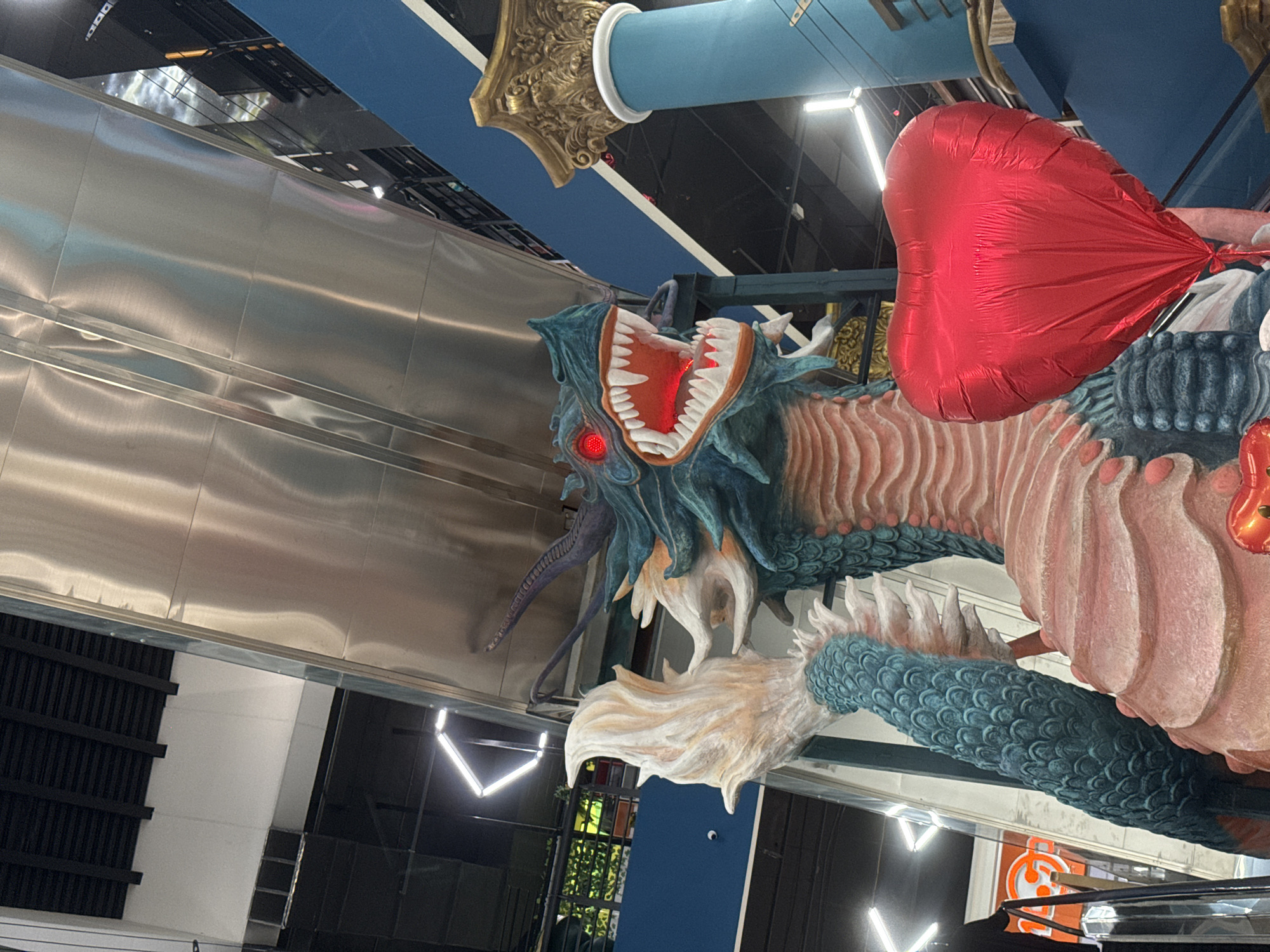 The image shows a large, decorative dragon sculpture with an open mouth displaying illuminated red interior, and red balloon at what seems to be a shopping center. The dragon has a teal body and a beige underbelly. A large red heart-shaped balloon is attached to the sculpture. The background includes a metallic wall, architectural details like columns and beams, and glimpses of the interior of the shopping center with lighting fixtures. Although the exact location within the city is uncertain from this image, the sculpture would most likely be at a shopping mall.