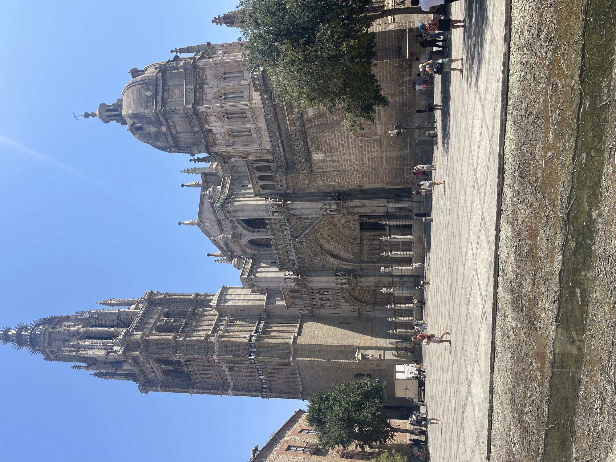 The image shows the exterior of the Toledo Cathedral in Toledo, Spain. The cathedral is a large Gothic structure with a tall bell tower on the left and a dome on the right. The facade features intricate stonework, statues, and arched doorways. In front of the cathedral is a large, paved plaza where several people are walking and standing. A portion of the plaza in the foreground appears to be dry and cracked, with some areas of murky water, possibly a remnant of a fountain or water feature. The sky is clear and blue.