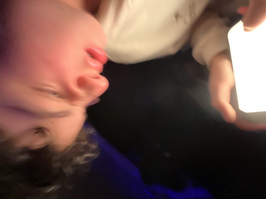 An individual is depicted in a motion-blurred, close-up image, located within the city of Santa Ana Acozautla, Mexico. The subject, a youth or young adult, is visible from an overhead angle, with the right side of their face, including an eye, nose, and a slightly open mouth, illuminated by a bright, rectangular object. Dark, curly hair is present on the left side of the head, and a reddish tint is noticeable on the right cheek. A light-colored garment, likely a sleeve, is visible on the lower right. Both hands hold a glowing, white rectangular electronic device, resembling a smartphone or small tablet, which acts as the primary light source for the scene. The individual appears to be looking downwards, engaged with the device. The background is largely dark and out of focus, with a subtle blue or purple light source apparent on the upper left.
