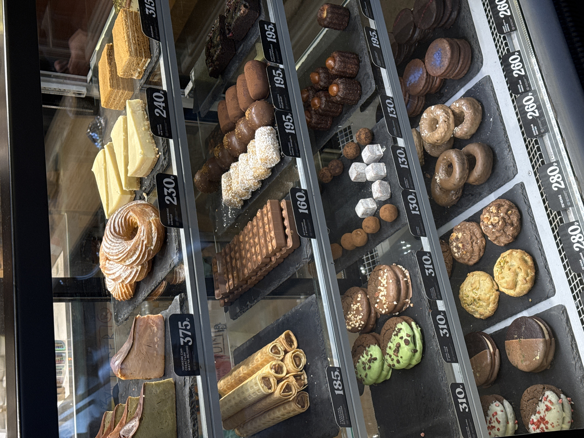 The image captures a vibrant display of various desserts and pastries inside a glass showcase, likely in a bakery or cafe in Moscow, Russia. The setting is indoors, with items neatly arranged on black slate trays across multiple shelves.
The display features a wide assortment of confections. On the left, slices of a multi-layered honey cake (Medovik, 315₽) and New York cheesecake (240₽) are visible, along with a large, powdered sugar-dusted pastry resembling a cottage cheese ring (Tvorozhnoe Koltso, 230₽). Further down, unique San Sebastian Matcha-Apple cheesecake (375₽) and stacked wafer rolls (Trubochka s varenyem, 185₽) are showcased.
The middle shelves feature numerous chocolate-based treats, including individual potato cakes (Kartoshka, 195₽), with variants like honey and coconut. Below these are chocolate-covered bars or biscuits (160₽).
Towards the right, more diverse items are displayed. There are small canelé-shaped pastries (195₽), alongside truffles and cuboid pieces of what appears to be Turkish delight (130₽). Further down, rows of small, glazed coffee donuts (Kofeynyy Ponchik, 260₽), various types of cookies (Pechenye, 280₽), and elaborate cookie sandwiches with fillings (310₽) are visible. Some cookies are dipped in chocolate, matcha green, or adorned with nuts and sprinkles.
Prices are clearly marked on black tags in rubles (₽) for each item or type of dessert, ranging from 130₽ to 375₽. Reflections of the store's interior, including parts of a person and the word "SURF," can be faintly seen on the glass of the display case. The overall impression is one of abundance and variety, typical of a specialty dessert shop.