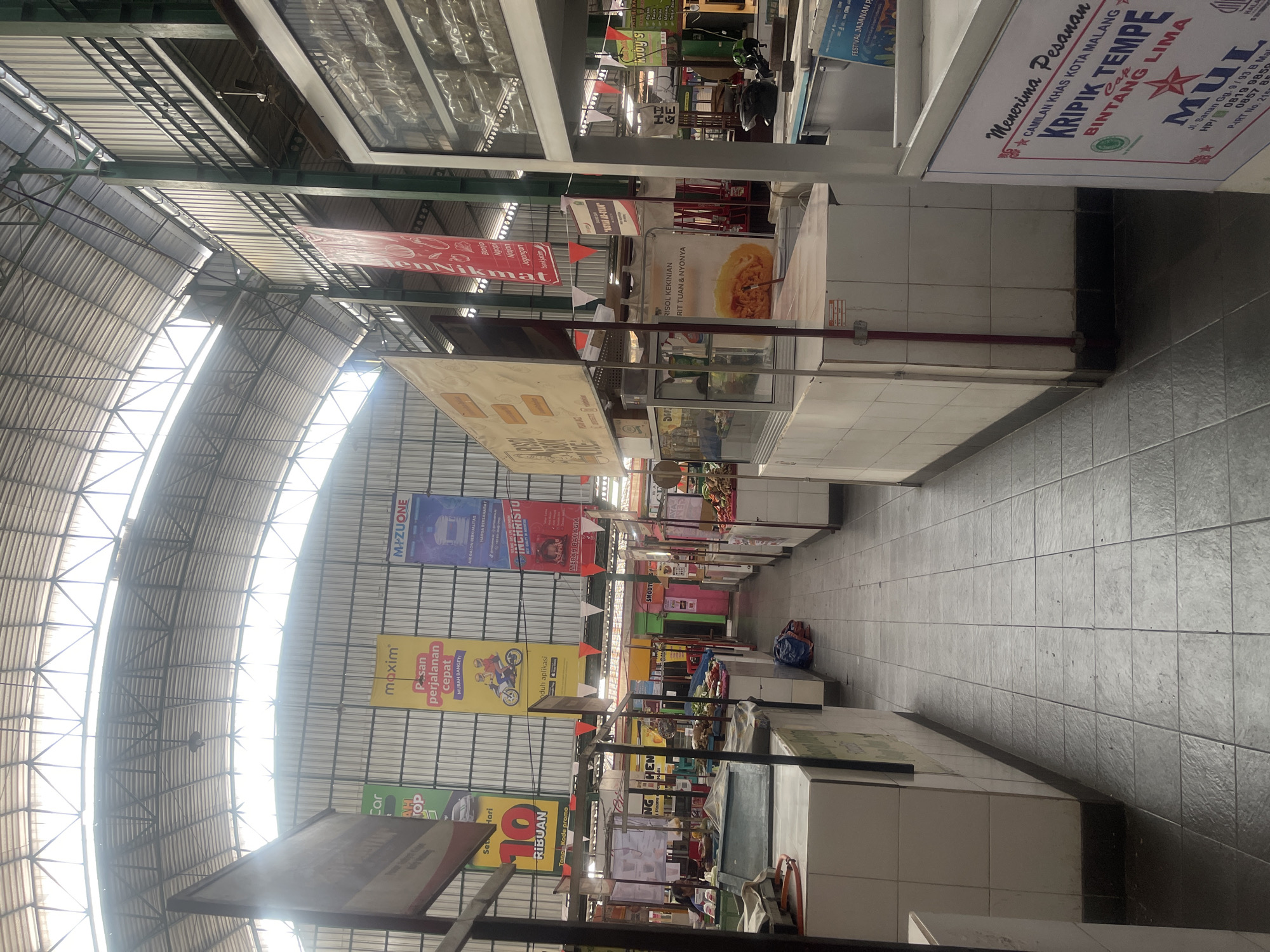 This image depicts the interior of a covered market in the city of Malang, Indonesia. The market features rows of stalls lining a tiled walkway. The stalls are primarily white with tiled countertops and some are covered with plastic sheeting. Various merchandise is visible, including produce, packaged goods, and what appears to be food items being prepared or displayed. Several signs and banners are hung throughout the market, some advertising products and others with promotional messages. The ceiling is constructed of metal beams and corrugated panels, with a skylight system allowing natural light to illuminate the space. One stall has a sign that reads "KRIPEK TEMPE BINTANG LIMA," indicating a food vendor selling fried tempeh chips. Another stall has a sign that says "RISOL KEKINIAN." The overall atmosphere suggests a typical Indonesian market environment.