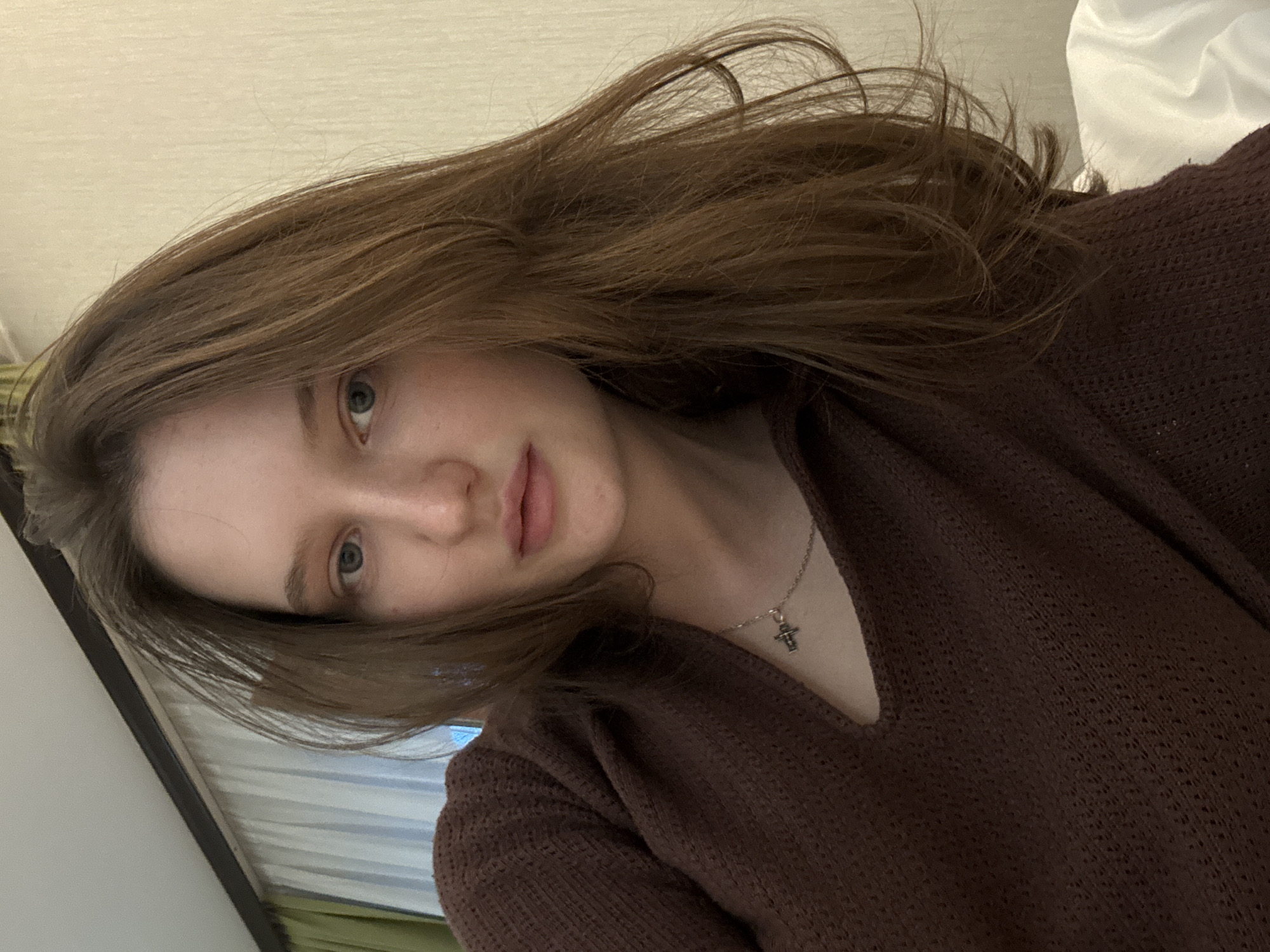 A medium shot captures a young woman from the chest up, facing the camera. She has fair skin, light blue eyes, and long, wavy, brown hair that frames her face and cascades over her shoulder. She is wearing a dark brown, textured sweater with a V-neck opening. A delicate silver necklace with a cross pendant rests on her collarbone. The background is a plain, light beige wall with a section of green and white curtains visible to the left. The lighting is soft and appears to be coming from an indoor source, possibly a lamp or natural light from a window. The overall impression is a casual, indoor portrait. The location is identified as the city of Журавлево (кемеровский), Russia.