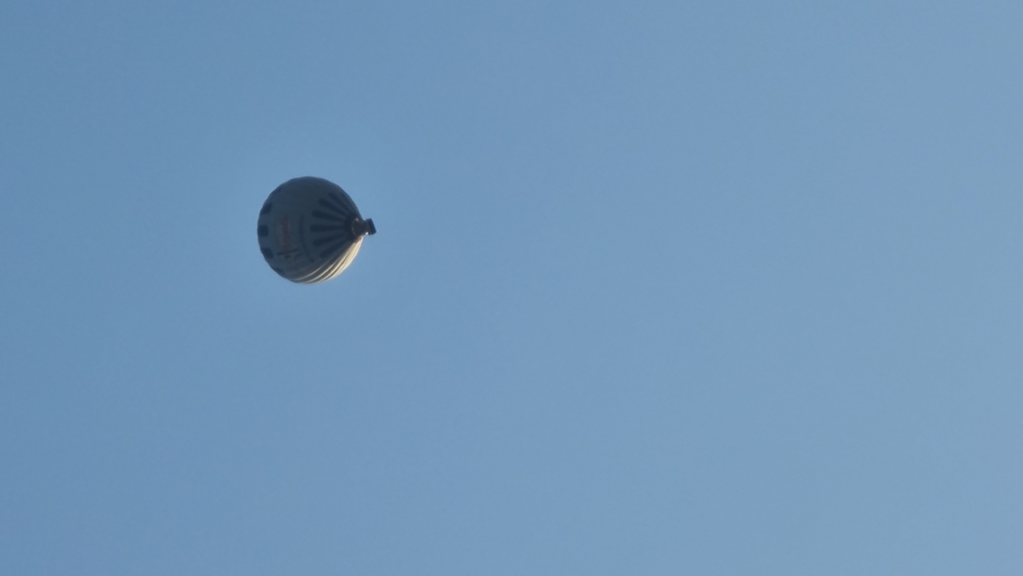 A hot air balloon is pictured against a clear blue sky. The balloon appears to be predominantly grey with some red accents and is illuminated from below, suggesting it is either lit from within or the sun is positioned low in the sky behind it. The balloon is in the upper right quadrant of the image. The scene is devoid of any other objects or people. The location is identified as Sant Joan Les Fonts, Spain, though no specific landmarks or context within the town are visible in the image.