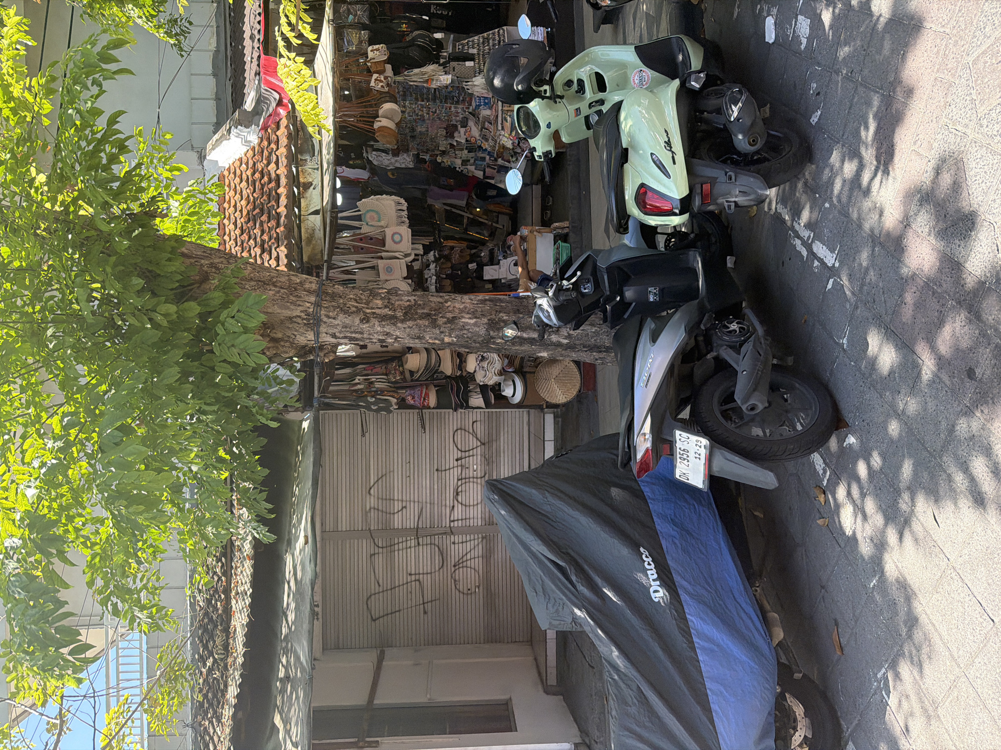 This image depicts a bustling street scene in Kuta, Indonesia, under bright daylight. In the foreground, several motorcycles are parked on a paved sidewalk. A prominent light green scooter, resembling a Vespa, has a black helmet resting on its seat. Adjacent to it is a gray/silver scooter, displaying a license plate "DK 2956 SC" with "12-29" (likely indicating registration month and year) printed below. Another vehicle, possibly a scooter, is partially covered by a blue tarp with the brand "Dracco" visible, and a black cover.
A large, leafy tree with a thick trunk stands prominently in the midground, casting shadows onto the ground and vehicles. Behind the tree, a vibrant storefront is visible, brimming with various goods. The shop appears to be a souvenir or general merchandise store, showcasing items like straw hats and intricately woven, fan-shaped bags. The building features a distinctive tiled roof.
In the background, a closed shop with a corrugated metal rolling door is present, marked with graffiti that appears to read "LONG JOHN" amidst other scribbles. Bright green foliage from trees and glimpses of other buildings or the sky complete the upper part of the scene. The clear, sunny weather is evident from the sharp shadows. The setting suggests a lively commercial area.