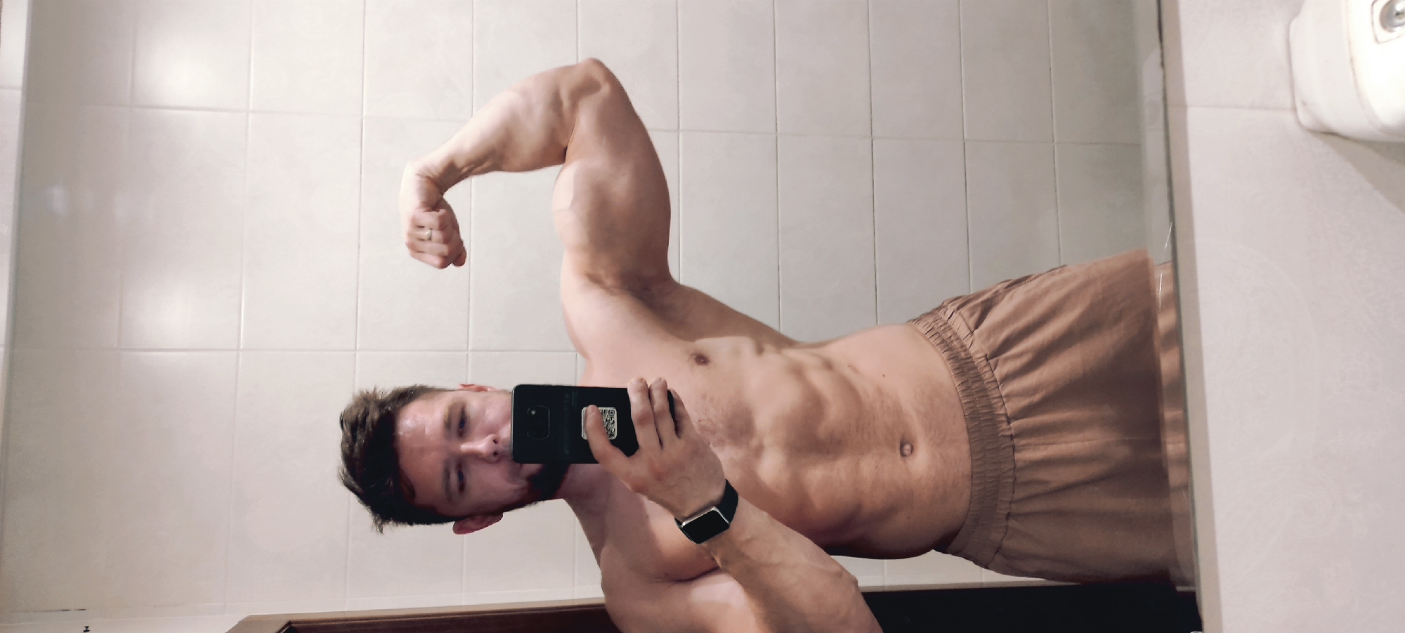 A muscular man is taking a selfie in a bathroom mirror. He is shirtless, wearing shorts, and flexing his bicep. He has a smartwatch on his wrist and is holding a smartphone. The bathroom has tiled walls and a hand soap dispenser. There is no information to indicate this photo was taken in the city of Fryazino, Russia.