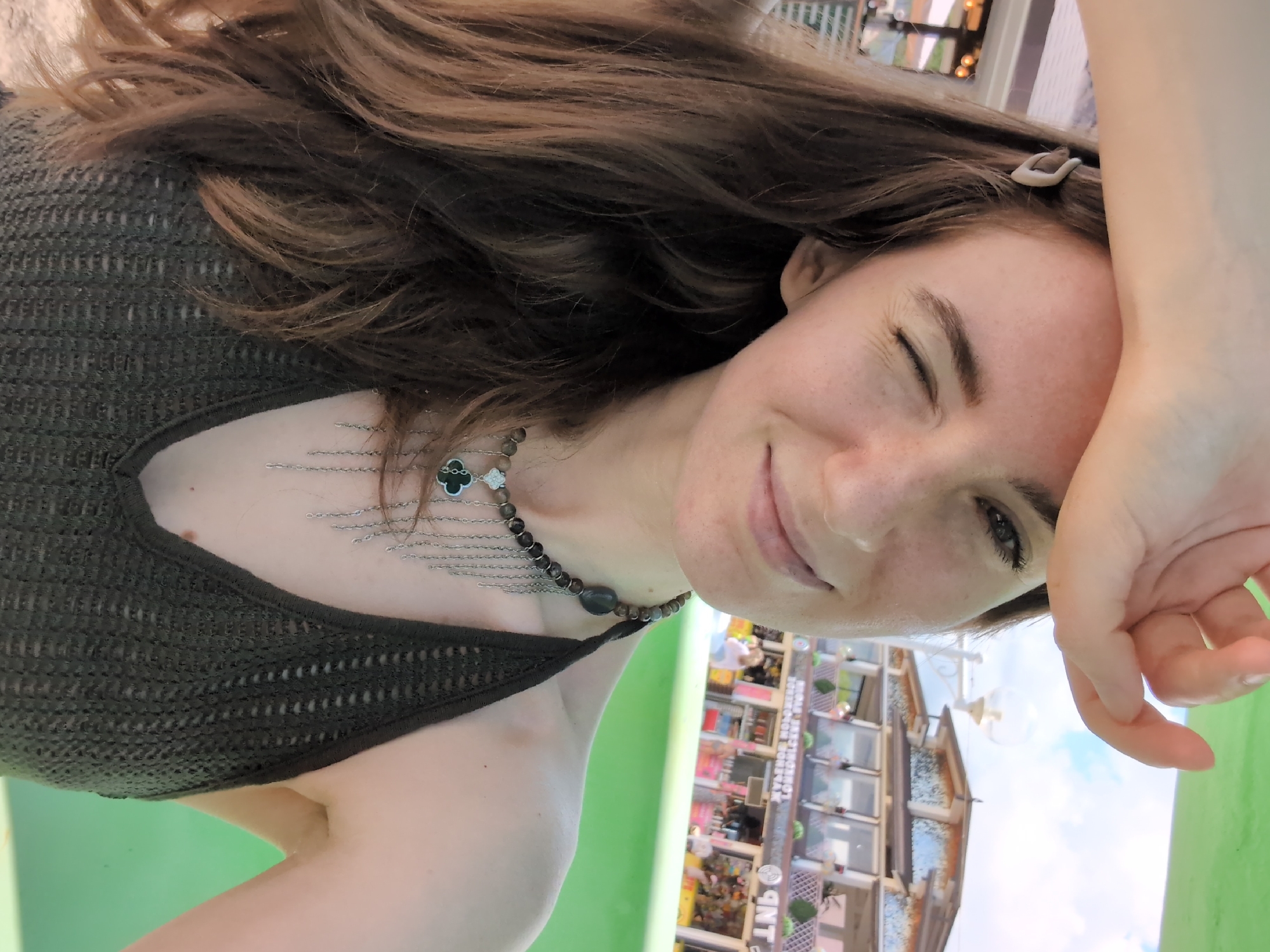 A young woman, facing the camera, winks as she shields her eyes from the sun with her right hand. She has long, brown hair, a light complexion with freckles, and is wearing a dark green, crochet-style halter top and a multi-strand necklace with a clover pendant. In the background, out of focus, are buildings that appear to be shops or restaurants, a green grassy area, and a bright blue sky with some clouds. The context suggests this photograph was taken outdoors, likely during the day, possibly in a tourist or recreational area. The location is identified as Divnomorskoye, Russia.