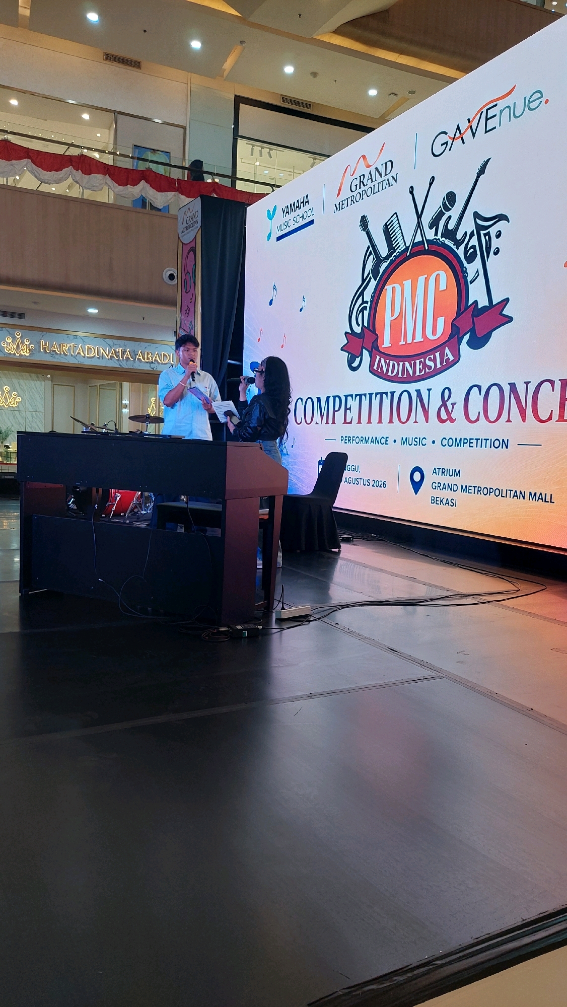 The image captures an indoor event stage setup in Bekasi, Indonesia, likely at the Grand Metropolitan Mall. The backdrop features a large LED screen displaying event branding, including "YAMAHA MUSIC SCHOOL," "GRAND METROPOLITAN," "GAVenue," and a prominent logo with "PMC INDONESIA." Text on the screen indicates "COMPETITION & CONCE," "PERFORMANCE • MUSIC • COMPETITION," "GGGU, AGUSTUS 2026," and location details: "ATRIUM GRAND METROPOLITAN MALL BEKASI."
In the foreground, a man in a light blue shirt is speaking into a microphone while holding a piece of paper. To his right, a woman in a black jacket is also holding a microphone and a paper. They appear to be interacting during a presentation or announcement.
In front of them is a large, dark-colored electronic keyboard or piano with musical instruments visible behind its front panel, including what looks like a drum. The stage floor is dark and reflective, with cables and a power strip visible. In the background, there is a mezzanine level with red and white draped decorations, and some storefronts or displays. The overall environment suggests a public event or performance space within a shopping mall.
