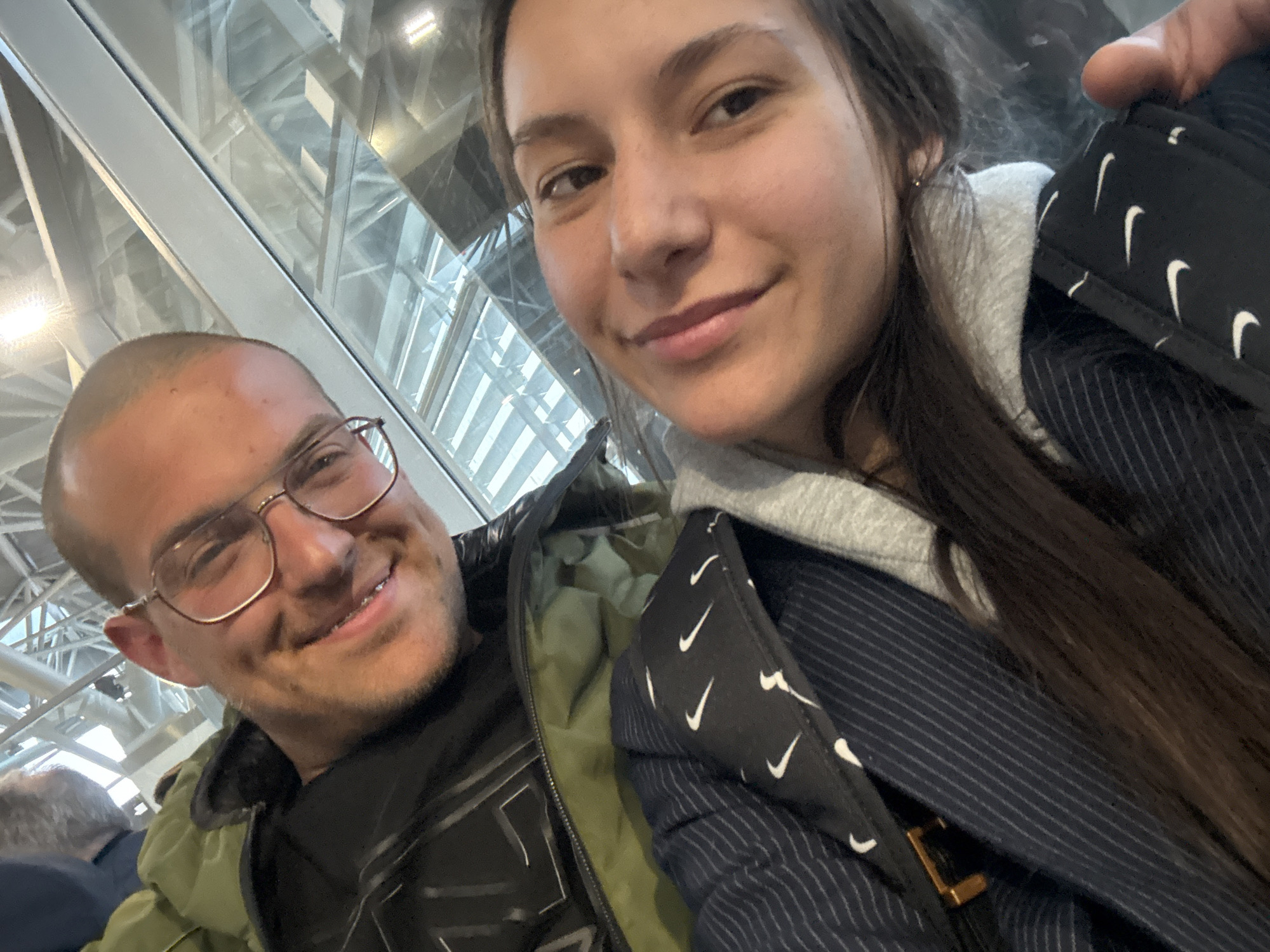 A selfie depicts a man and a woman. The man, positioned on the left, has a shaved head, glasses, and is wearing a green jacket over a black shirt. He is smiling. The woman, on the right, has long brown hair and is wearing a gray hoodie underneath a pinstripe jacket. Part of a black bag with a repeated white checkmark pattern is visible. Behind them, a window reveals an indoor structure with a geometric framework. The location appears to be an airport or train station based on the architectural elements seen through the window. While the image contains clues resembling modern transportation hubs, there is insufficient information within the picture to positively identify the location as Bratislava, Slovakia.