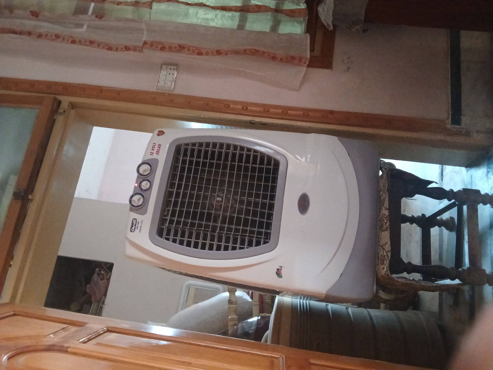 An outdoor air cooler is displayed in the foreground, placed on a wooden stool. The cooler is white and grey, with a large grill at the front. To the left of the cooler, a portion of a wooden door frame is visible. The background shows a room with a window covered by a light-colored curtain with a decorative pattern. A light switch is mounted on the wall to the right of the door frame. The environment appears to be indoors, likely a residential space within Peshawar City Tehsil, Pakistan, judging by the architectural and decorative elements.