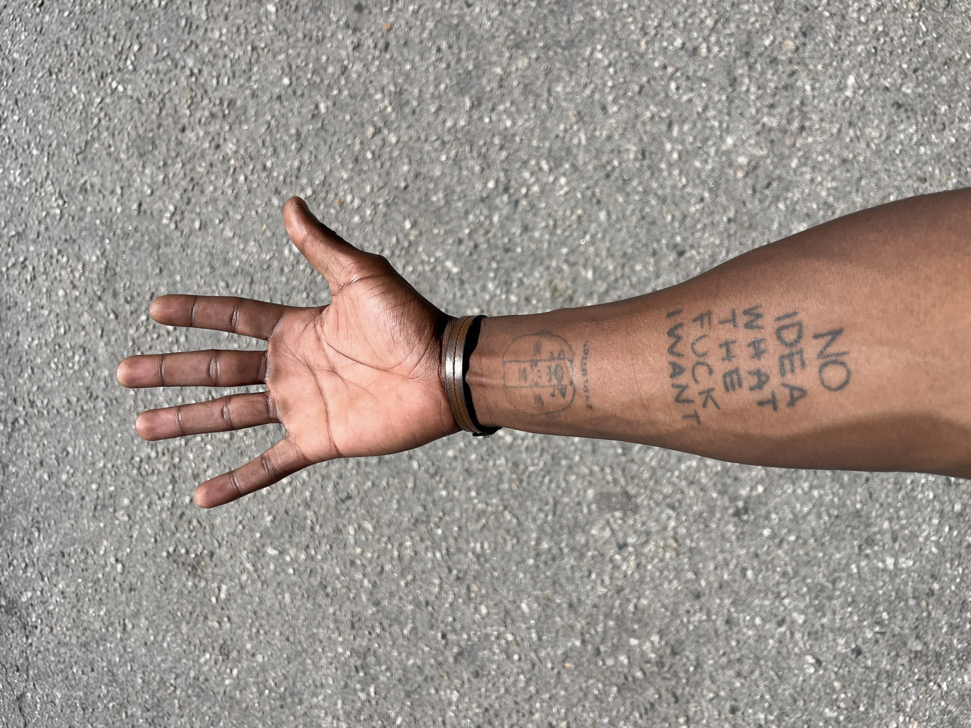 An arm and hand are extended against an asphalt surface. The person has brown skin and several tattoos. One tattoo reads "NO IDEA WHAT THE FUCK I WANT." There is a circular tattoo closer to the wrist, and a leather wristband. The setting is nondescript and does not indicate a location in Barcelona, Spain.