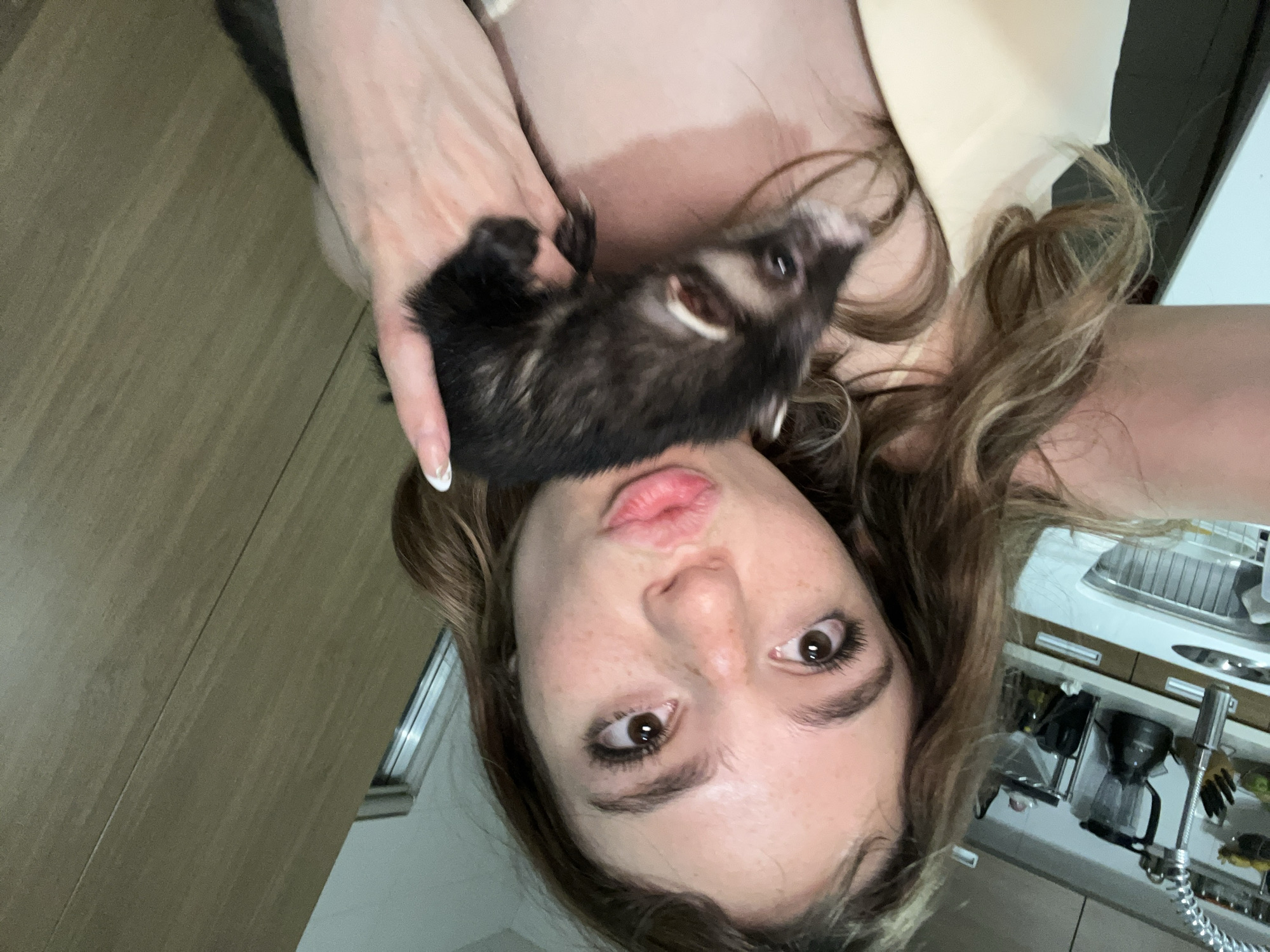 A woman is holding a ferret close to her face in what appears to be a kitchen. The woman has light skin, brown hair, and brown eyes. She is wearing a light-colored top. The ferret is black and white. The kitchen is modern and appears to have light wood cabinets and stainless steel appliances. It is difficult to discern the location within San Bernardino Tlaxcalancingo, Mexico, due to the limited visual information.