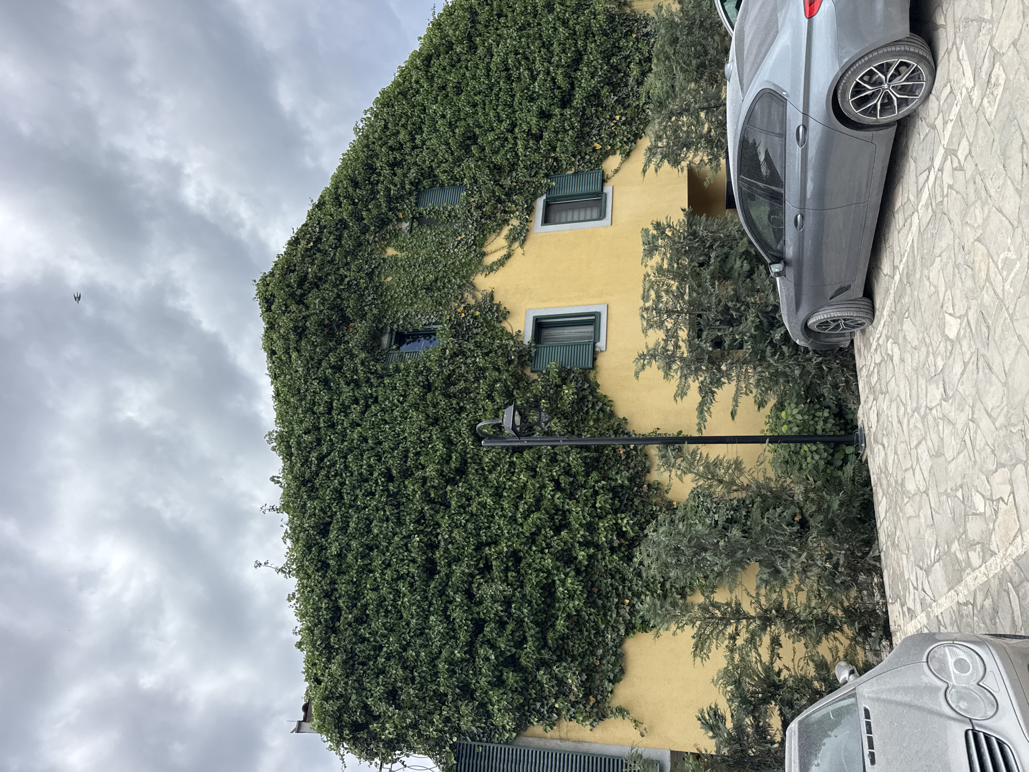 A yellow, ivy-covered building with green shutters is the primary focus of the image. Two grey cars are parked alongside the building. A black lamp post is visible in front of the building. The sky is cloudy. The ground is paved with light-colored stones. The location is Lastva Grbaljska, Montenegro. No people are visible.