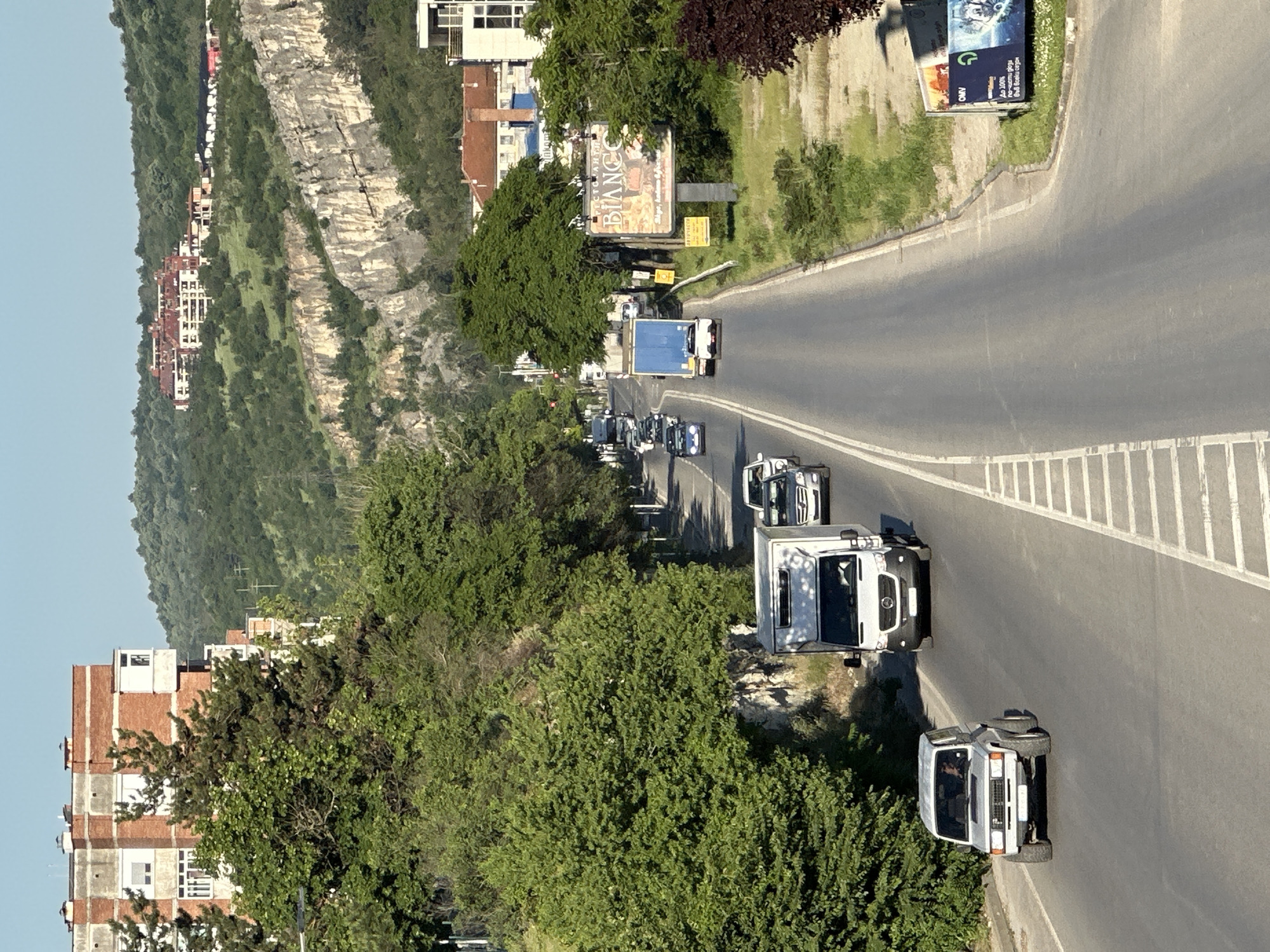 The image shows a street scene in Veliko Tarnovo, Bulgaria, featuring a road with several vehicles, including trucks and cars. On the left side of the road, there are buildings and a cliff face. Vegetation, including trees and bushes, are also present along the roadside. Billboards are visible on the right side of the road.