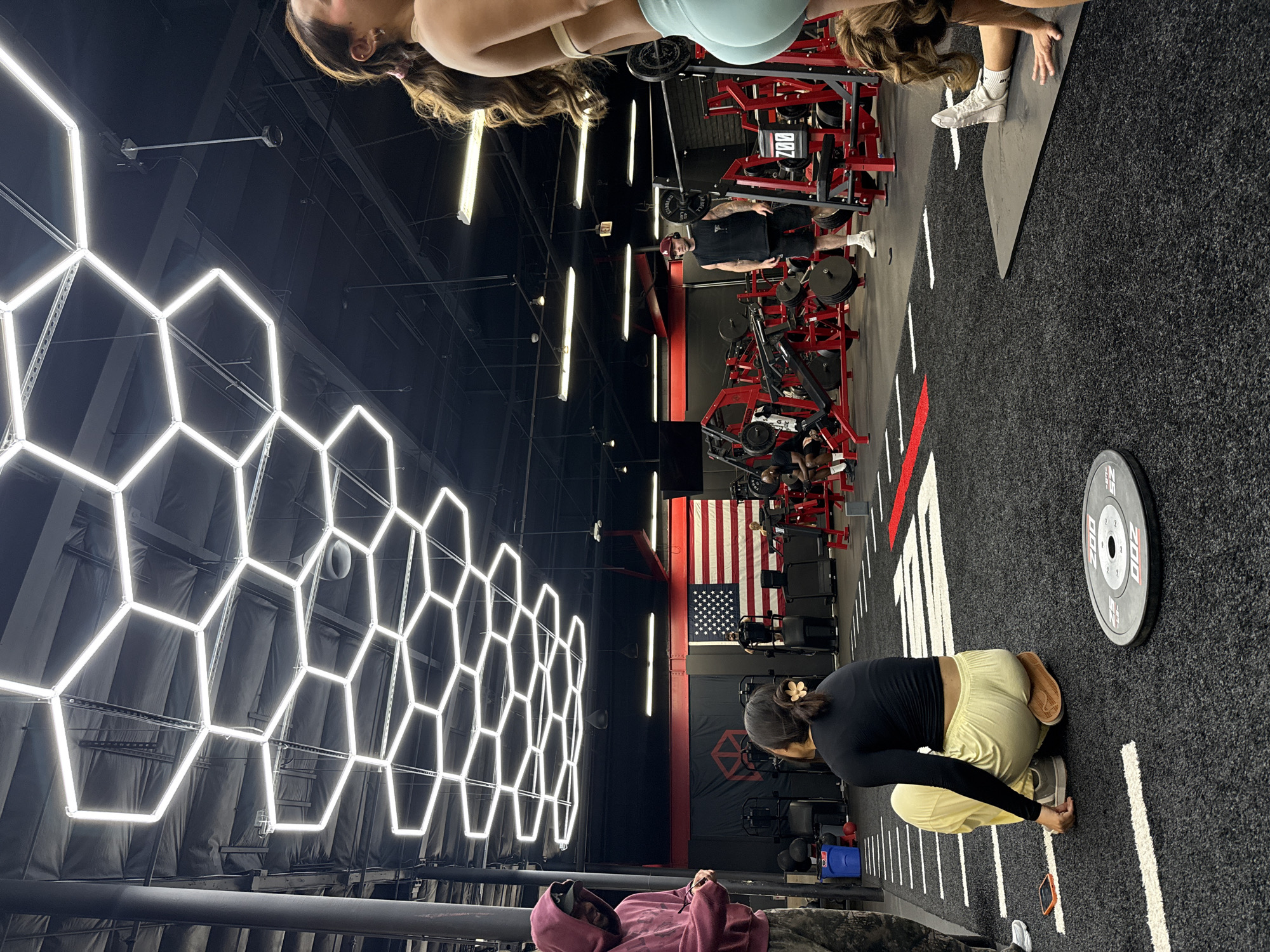 The image depicts an indoor scene within a modern gym or fitness center, likely located in Los Angeles, USA. The perspective is tilted, showing various individuals engaged in workouts amidst specialized equipment.
In the foreground, a woman in a black long-sleeve top and light yellow pants crouches on a dark, carpeted floor marked with white and red lines. Her hair is tied in a bun with a flower clip. Near her, a grey 25-pound weight plate is visible on the floor, labeled "25 LBS" and "200". A smartphone also rests on the floor beside her. To the left, another person in a maroon hoodie is partially visible, facing away.
Further into the gym, several people are present. A woman with long, brown hair, wearing light-colored athletic wear, is seen lying on a mat. Nearby, a man in a black tank top, dark shorts, and a red beanie stands among multiple red weight machines. Tattoos are visible on his arms. Text like "200" and "CH" can be seen on some of the red equipment. Other indistinct figures are visible around more gym machines in the background.
The gym features a modern design with high ceilings, dark walls, and a distinctive lighting arrangement. A large array of bright, white hexagonal LED lights forms a geometric pattern on the left wall. Rectangular fluorescent lights are also visible on the ceiling. On a far wall, a large American flag is displayed, and beneath it, a black banner with a red geometric logo is hanging. The overall setting is well-lit by artificial sources, suggesting an active gym environment.
