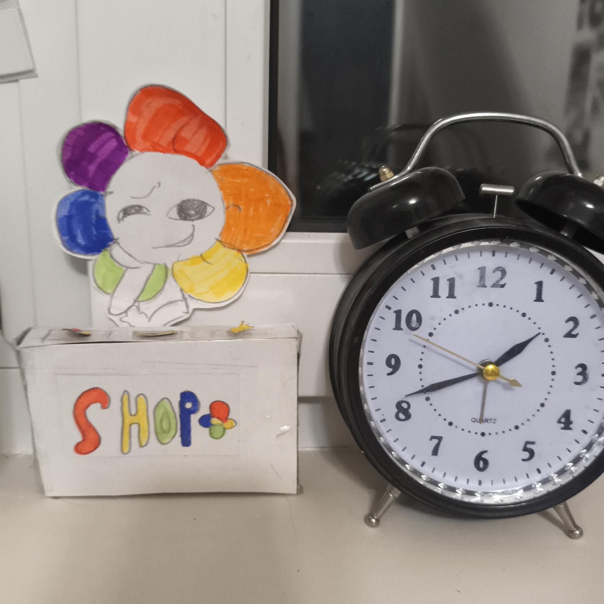The image shows a white surface with a black alarm clock on the right, displaying 2:42. Next to it is a small white box with "SHOP" written on it in colorful letters. On top of the box is a colorful, hand-drawn flower-like character with a winking face. The background consists of a white wall and a dark window. The location and city are unknown.