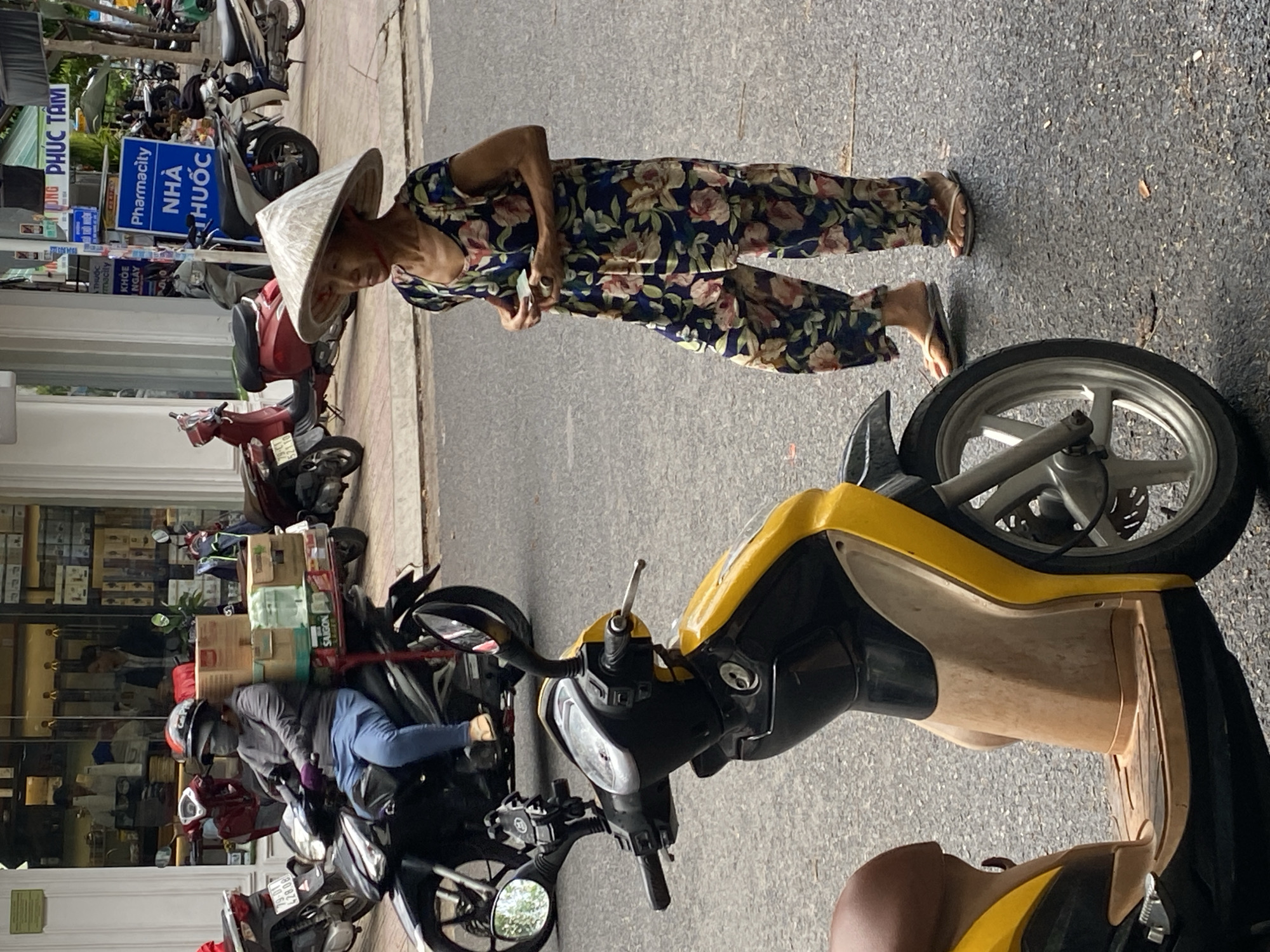 This media file captures a street scene in Nha Trang, Vietnam, likely during daytime, given the bright lighting. The primary subjects are two individuals, each associated with a motorbike.
In the foreground, a woman wearing a traditional conical hat and a floral-patterned dress is walking on the asphalt road. She is holding a small item in her hand, possibly a receipt or a piece of paper, and appears to be looking down. Her posture and the slight tilt of her head suggest a moment of focus or contemplation. She is wearing flip-flops.
Further back and to the left, a person is seated on a motorbike, wearing a helmet and a grey long-sleeved top with blue pants. They appear to be wearing gloves. This motorbike is laden with boxes and other items stacked on a rack behind the rider, indicating a delivery or transportation of goods. The background shows a row of shops with numerous shelves stocked with products, suggesting a commercial area. Several other motorbikes are parked or moving in the vicinity, typical of the bustling street life in Vietnamese cities.
Visible text includes signs with Vietnamese characters. One prominent blue sign reads "Pharmacy NHÀ THUỐC HIỂU PHUOC." Another sign near it displays "PHARMACITY" in English. The overall impression is a snapshot of everyday life, commerce, and transportation on a busy street in Vietnam.