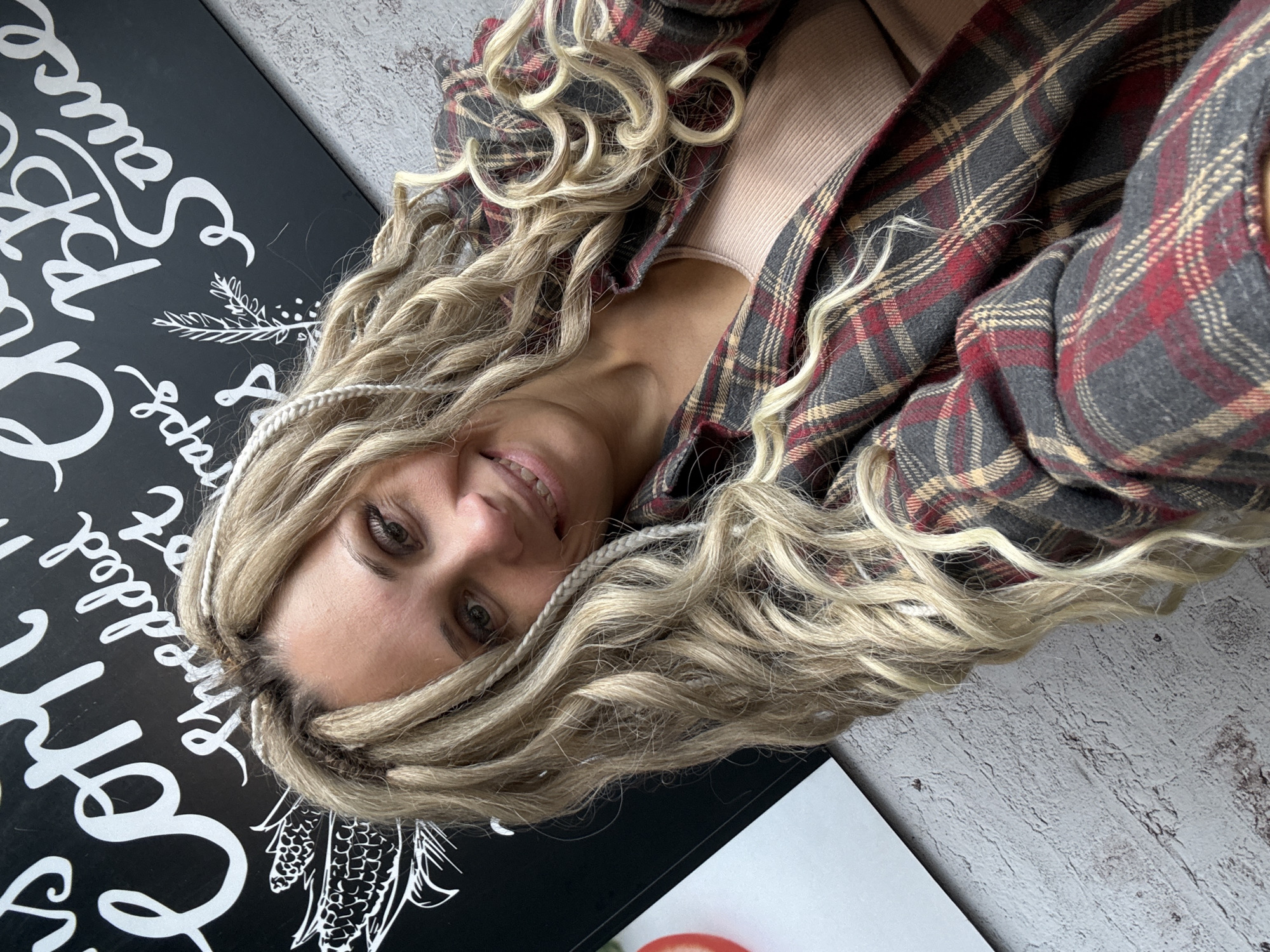 A woman with blonde dreadlocks and a plaid shirt lies on a gray, weathered surface. She is smiling at the camera. To her left is a black sign with white lettering and illustrations of plants. A second sign, mostly white, is visible on the lower right corner. The location is not discernible from this image.