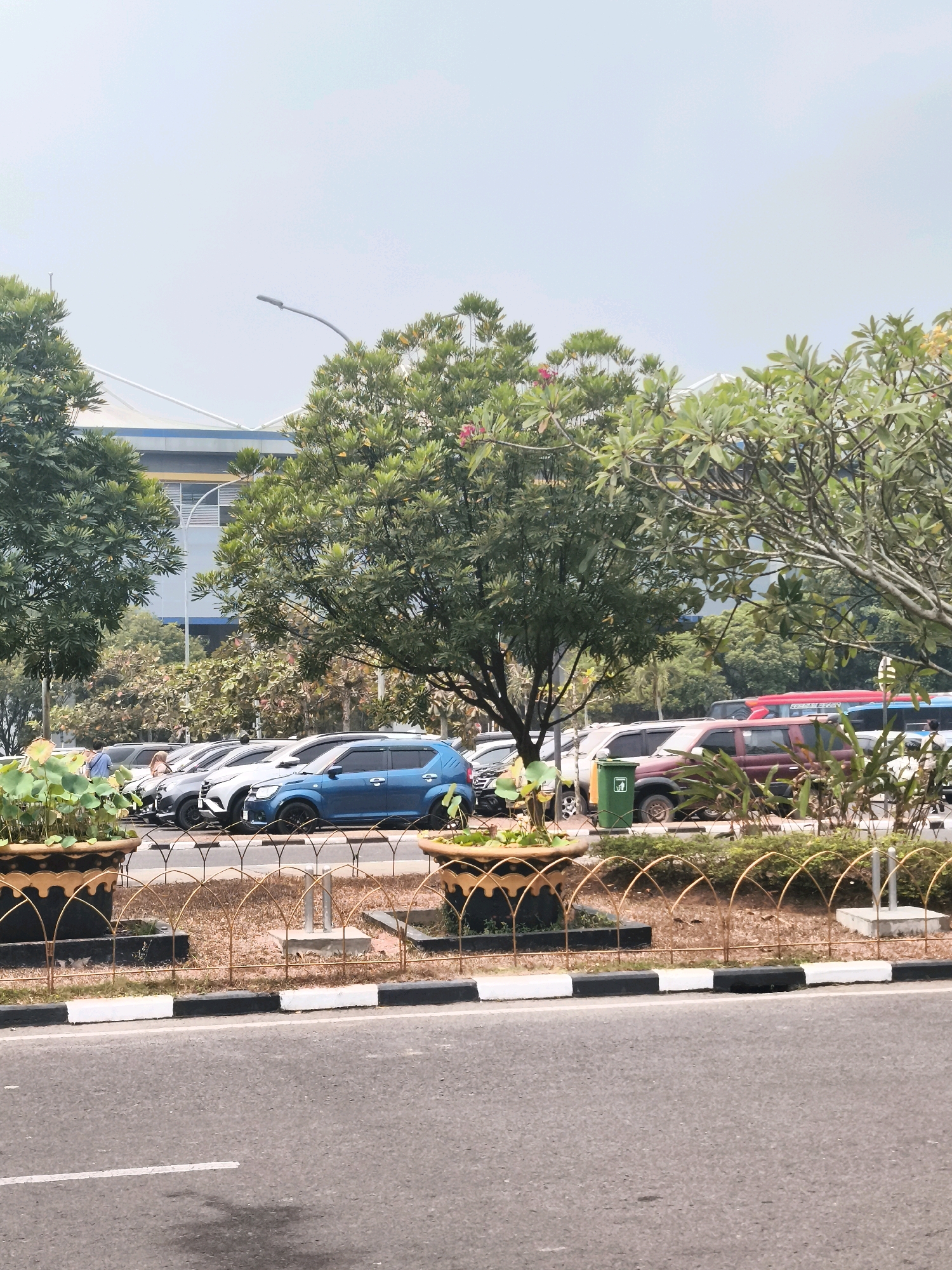 An outdoor, daylight, medium shot captures a parking lot scene in Palembang, Indonesia. In the foreground, a paved road with a white line is visible. Behind the road, a decorative metal fence with an arched design encloses a landscaped area. Two large, decorative planters with gold trim and dark interiors are positioned at the ends of the fence.
Beyond the fence, a line of parked cars stretches across the frame. A bright blue SUV is prominently featured in the middle of the car line. Other vehicles, including a maroon SUV and parts of a red bus and a blue bus, are also visible. A green trash bin is located next to the maroon SUV.
In the background, lush green trees with some pink flowers are abundant, framing the scene. Behind the trees, a modern building with blue and white accents is partially visible. A street light with a curved arm extends from behind the trees, reaching towards the sky. The sky is a pale blue with a slight haze.