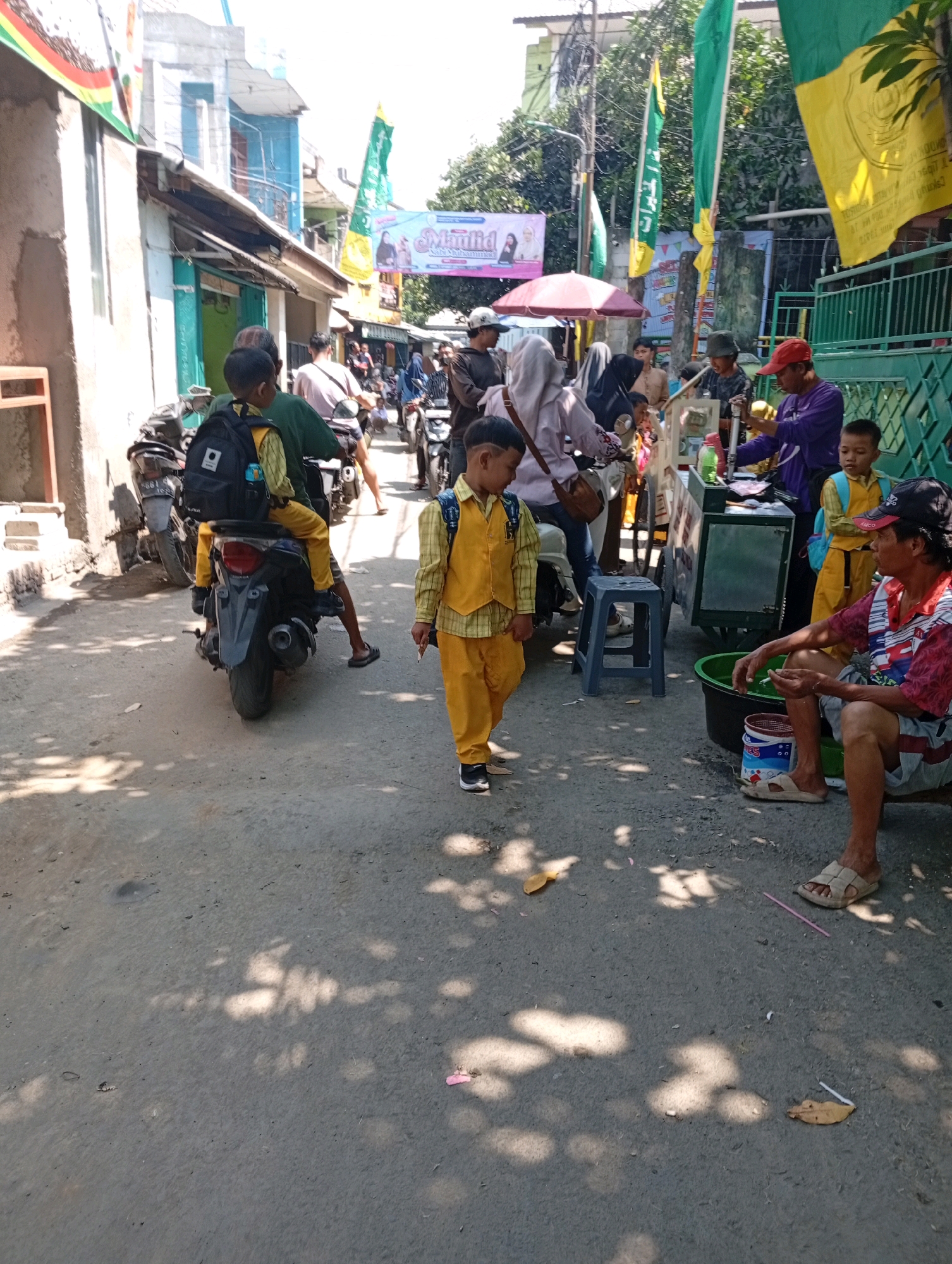 A street scene in East Jakarta, Indonesia, features a diverse group of people and vehicles. In the foreground, a young boy in a yellow uniform walks along the dusty, uneven road, casting a shadow from the dappled sunlight filtering through trees overhead. He is followed by a man on a motorcycle, also dressed in yellow. Behind them, a woman in a pink headscarf stands near a food cart, engaged in conversation with a vendor wearing a purple shirt and red cap. Several other individuals, including children and adults, are scattered throughout the scene, some on motorcycles, others walking or standing near shops and stalls. Flags with green and yellow colors are visible, suggesting a local event or celebration. The buildings lining the street are simple, some appearing to be single-story structures with weathered paint. The overall impression is one of a bustling urban environment with everyday activities taking place.