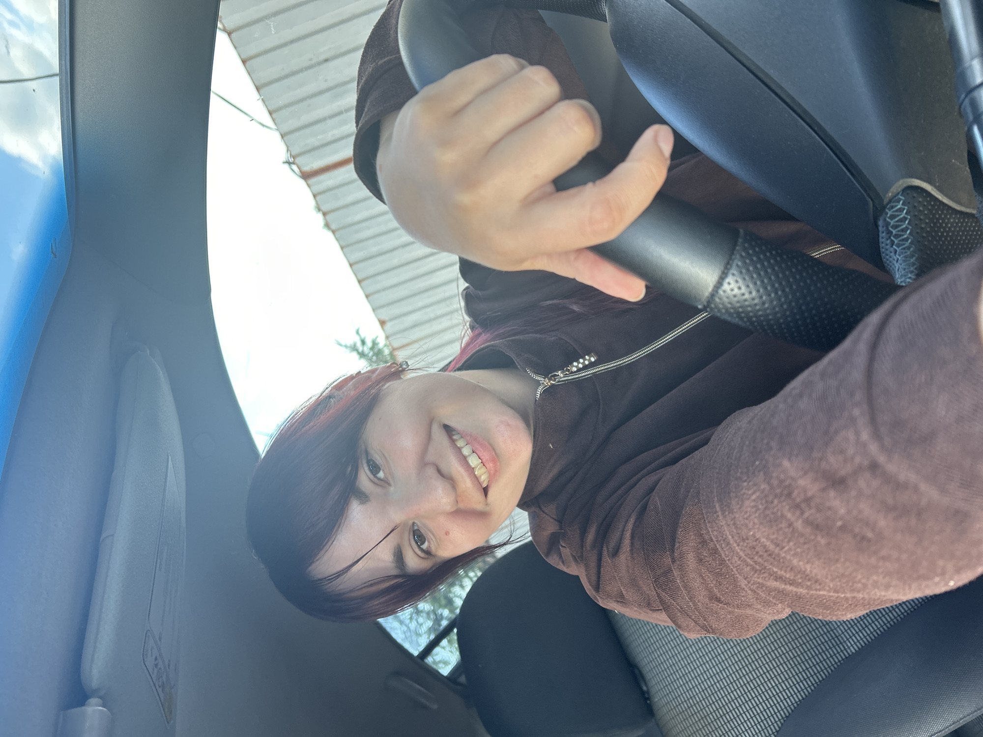 The media file shows a person in the driver's seat of a car. The person, a woman with dark hair and bangs, is smiling and looking at the camera. She is wearing a brown hooded jacket. Her hands are on the steering wheel. The interior of the car is visible, including the steering wheel, dashboard, and a sun visor. The sun visor has some text on it, but it is not legible. The scene appears to be outdoors, as indicated by the sunlight and the trees visible through the car windows. The time of day appears to be daytime, and the weather seems to be clear. The location is given as Фоки, Russia. The overall mood is cheerful and casual.