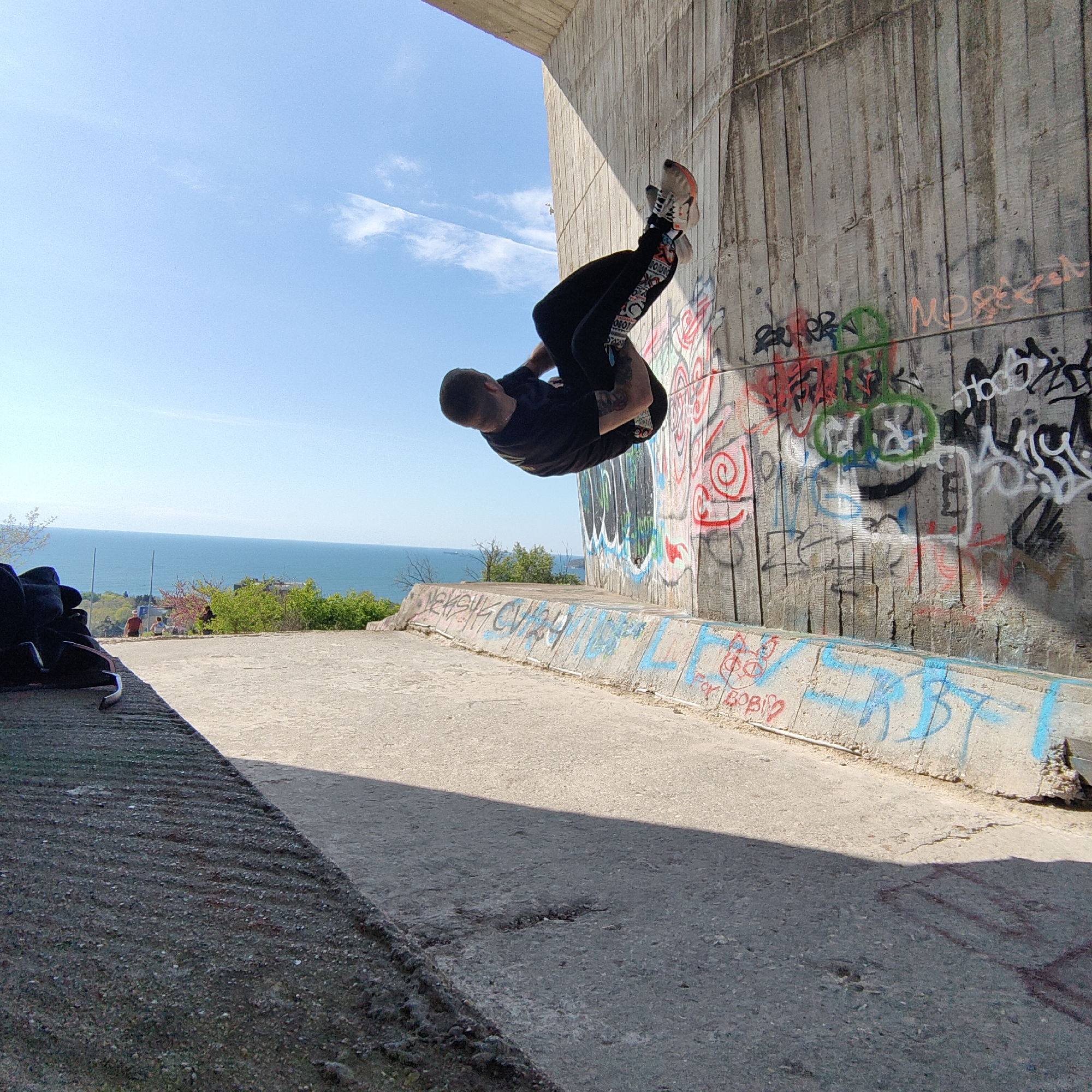 The image captures a man in mid-air, performing a gymnastic or parkour move against a graffiti-covered concrete wall. The location is likely Varna, Bulgaria, based on the ocean visible in the background. The man is wearing a black shirt, dark pants with printed designs, and shoes. He appears to be executing a backflip or similar maneuver. The wall is heavily adorned with various graffiti designs, including writing and abstract patterns. The ground is concrete, with a shadow cast across the surface. In the background, the Black Sea meets the blue sky at the horizon line. There are trees and some visible structures along the coastline. The lighting suggests it is daytime, with a clear sky.