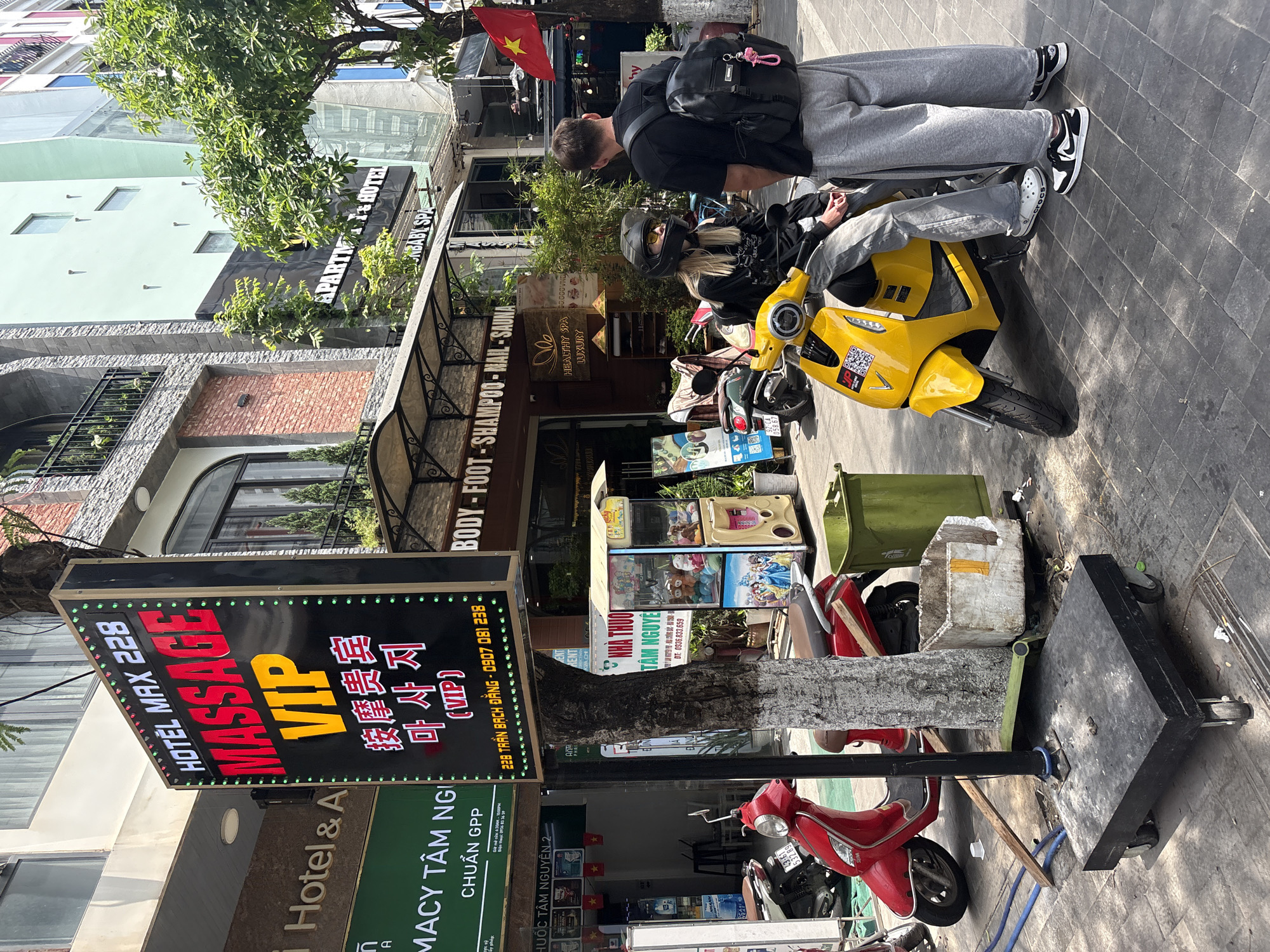 A street scene in Đà Nẵng, Vietnam, features a yellow scooter with a helmeted rider. A man with a backpack stands next to the scooter. The environment includes a massage parlor sign, a pharmacy sign, shops, scooters, and various street objects, creating a busy urban atmosphere. The signs feature Vietnamese and Korean characters.