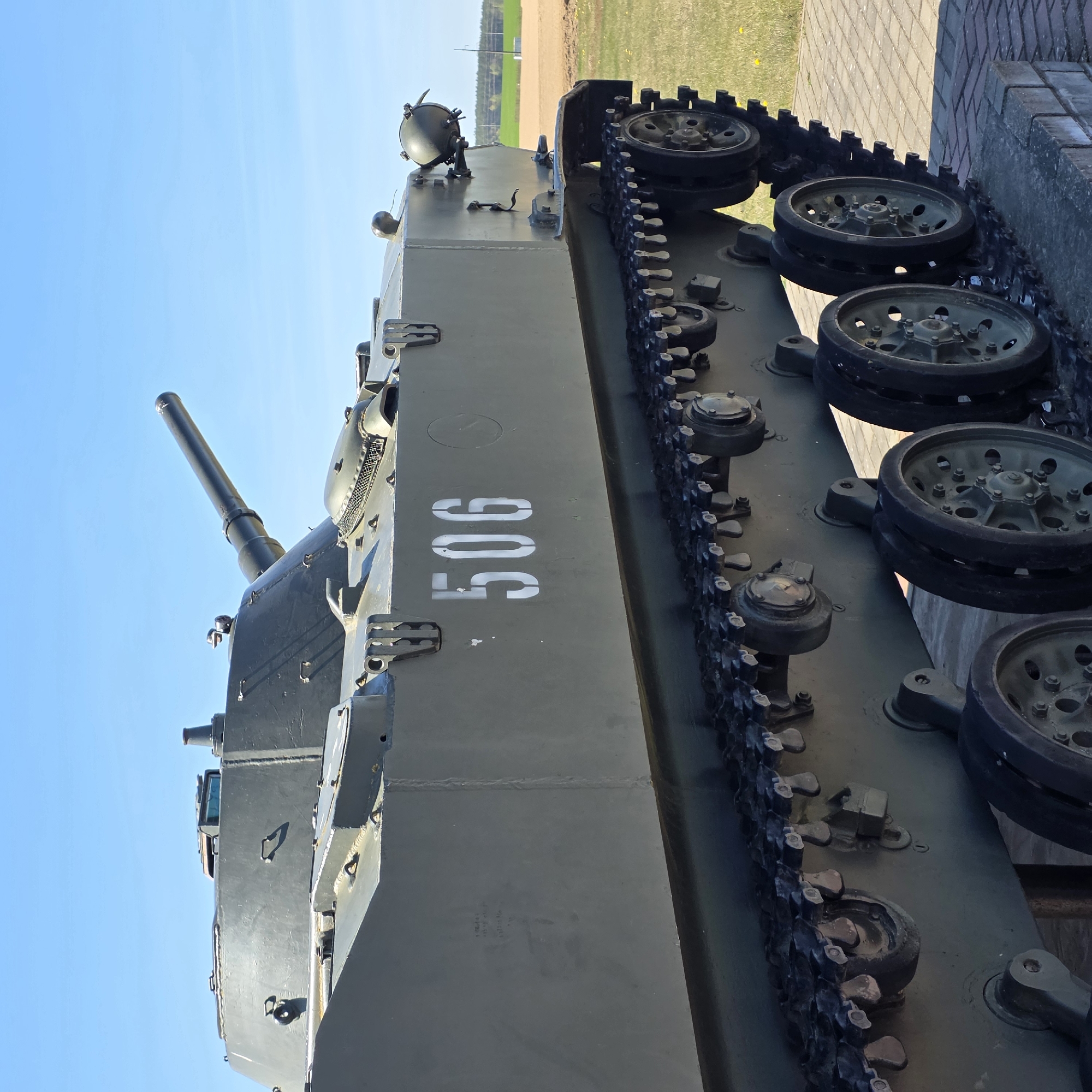 The image shows a military tank. The tank is painted in an olive drab color. The number "506" is stenciled on the side of the tank in white. The tank is parked on a paved area, adjacent to a grass field and a brick wall. The location is in Свіслацкі сельскі Савет, Belarus.