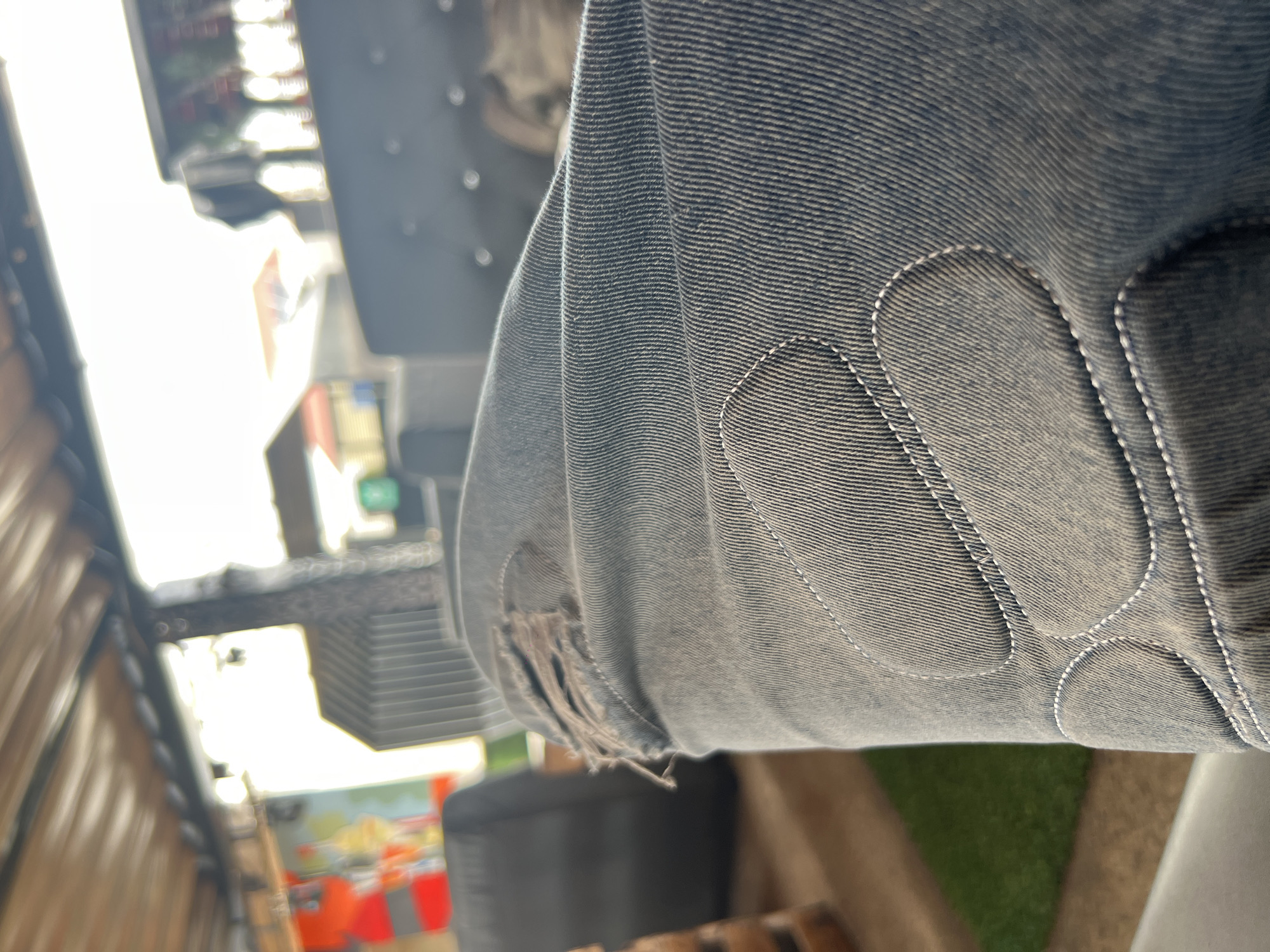The image shows a close-up view of denim jeans with a ripped knee. The jeans are a light blue-grey color and have visible stitching details, including decorative stitching around reinforced patches. In the background, out of focus, is an outdoor seating area with what appears to be a thatched roof overhead. Some furniture, possibly couches or chairs, is visible, along with a patch of artificial grass. The overall impression is of a casual, possibly urban outdoor setting. There is no specific indication of the location being in Howayi, Nigeria.