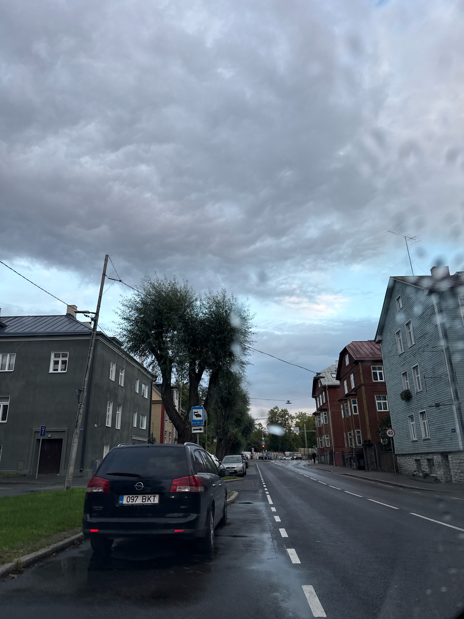 The image depicts a street scene in Tallinn, Estonia, under an overcast sky. The wet asphalt road reflects the surrounding buildings and sky, suggesting recent rain. On the left side of the street, a dark Opel Vectra is parked by the curb. Behind it, a gray, multi-story building with white-framed windows stands. Across the street, a row of traditional European buildings lines the road, including a tall, light blue wooden house with multiple windows and a red-roofed building. A speed limit sign indicating 40 km/h is visible on the right side of the road. The street is mostly empty, with a few cars visible in the distance and a lone pedestrian walking on the sidewalk. Tall trees with dense foliage are present along the street, adding to the urban landscape.